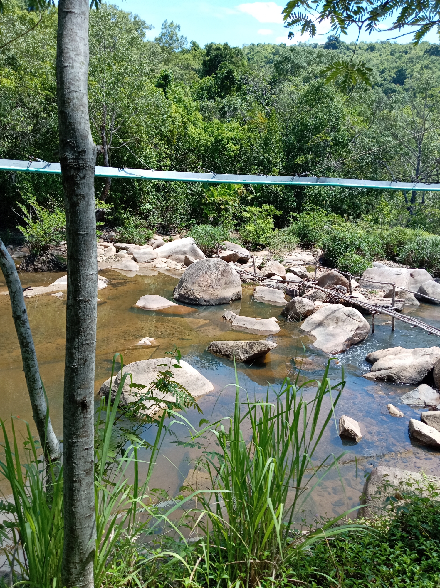 A river flows through a rocky landscape in Nam Ninh Hoa, Vietnam. The water is shallow and clear, revealing the riverbed below. Large, smooth rocks are scattered throughout the river, some partially submerged. A simple wooden bridge crosses the river, constructed from branches and logs. Dense green foliage and trees line the riverbanks, creating a lush and natural environment. The sky above is blue with a few wispy clouds. In the foreground, tall green grass grows along the edge of the water. A metal pipe or conduit runs horizontally across the upper portion of the image, supported by posts, suggesting some form of infrastructure in the area.