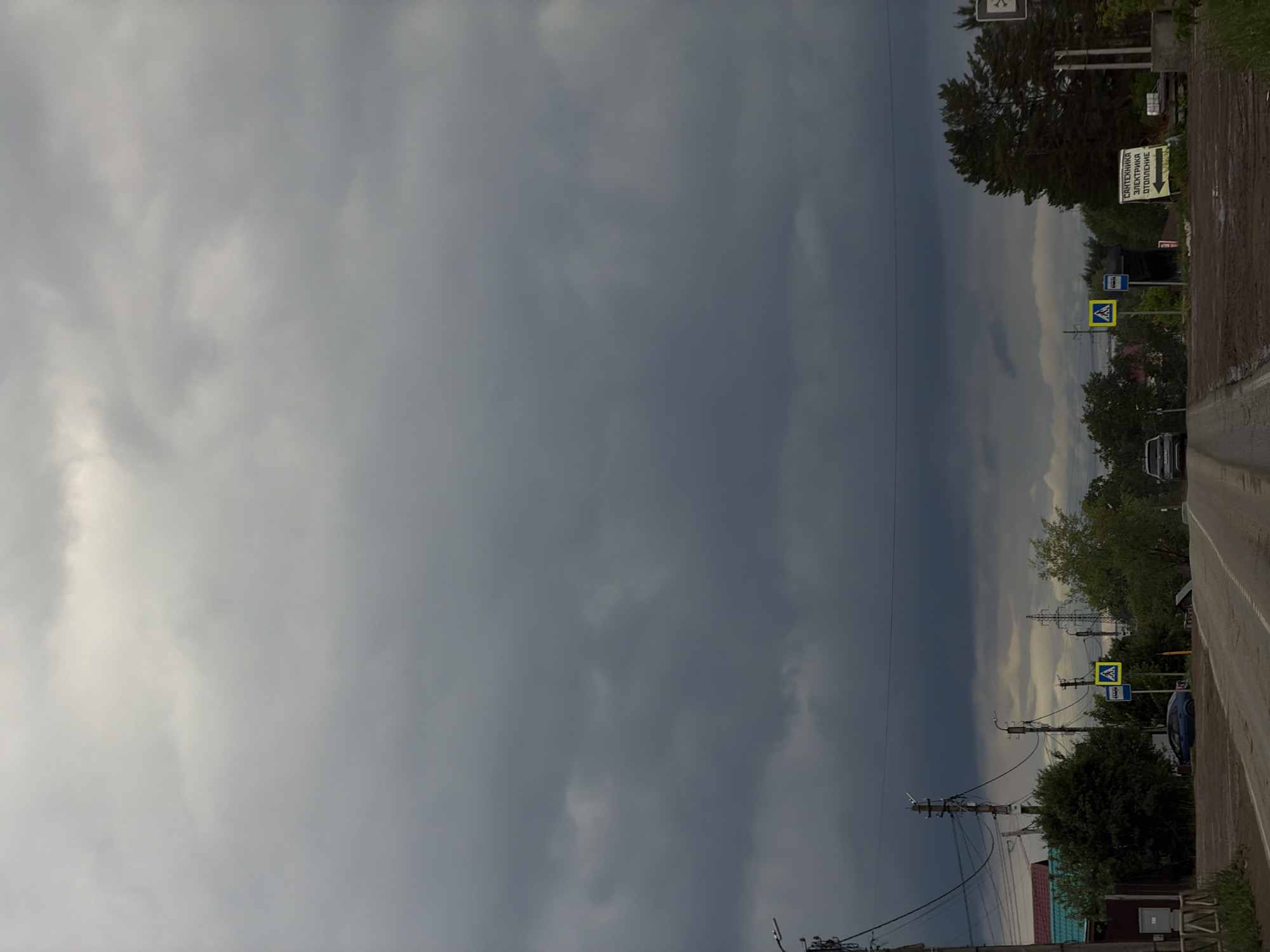 The image shows an overcast sky above a road in Chaikovsky, Russia. The road runs vertically, with a dark, stormy sky taking up the majority of the left side of the frame. Power lines and utility poles are visible along the right side of the road. Traffic signs, including pedestrian crossing signs, and a bus stop sign are present on the roadside. Buildings and foliage can be seen beyond the utility poles. Several cars are parked or traveling on the road.