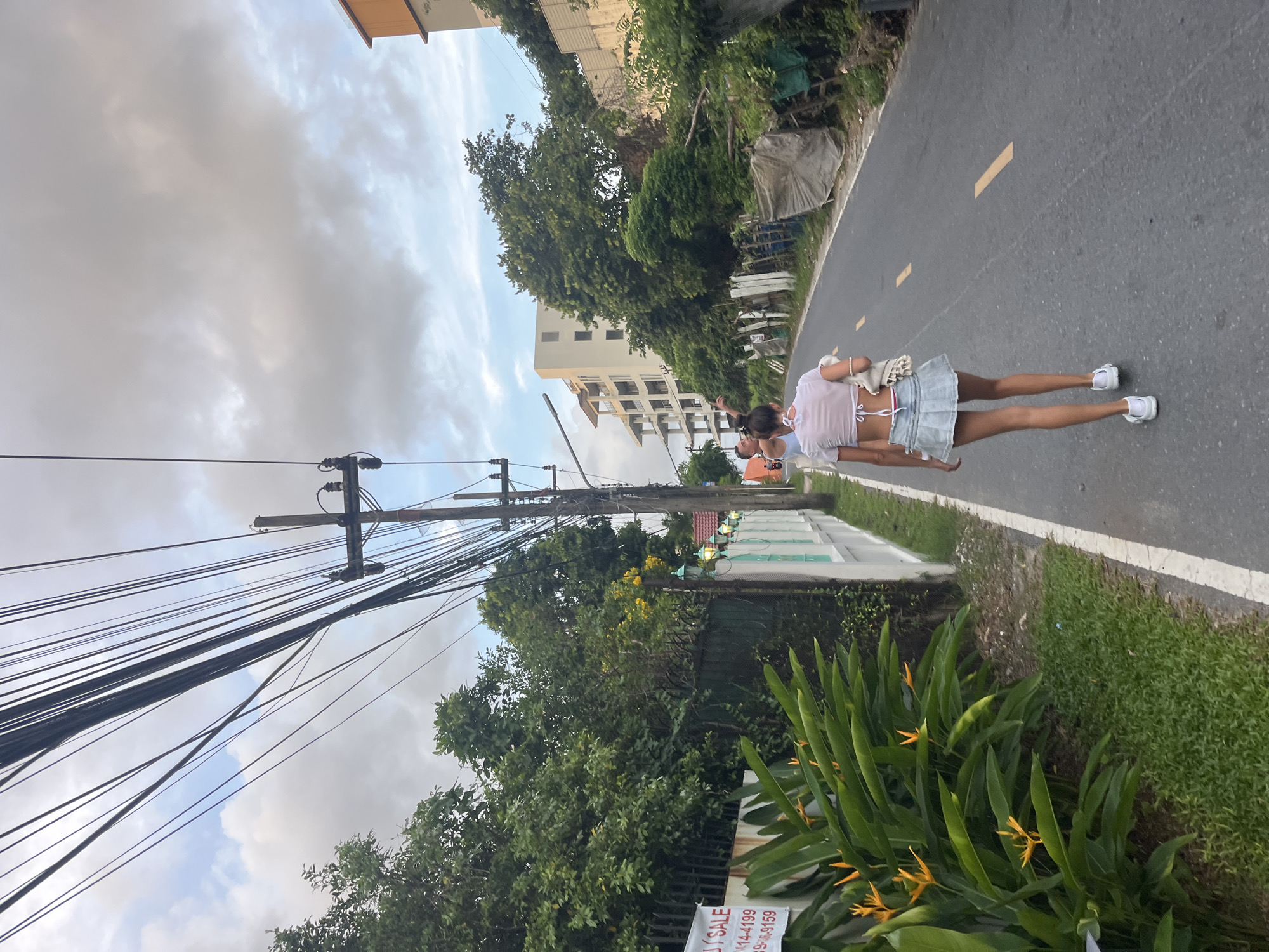 The image depicts a street scene in Choeng Thale, Thailand, characterized by a paved road, overhead power lines, lush greenery, and buildings. In the foreground, a woman with dark hair is walking away from the camera, wearing a white crop top and a light blue denim skirt. She has white clogs on her feet and carries a cream-colored bag. To her left, there are large green leaves with yellow flowers, and a white fence runs alongside the road, adorned with decorative lamps. Further down the street, a multi-story building is visible, partially obscured by trees. The sky is overcast with clouds.