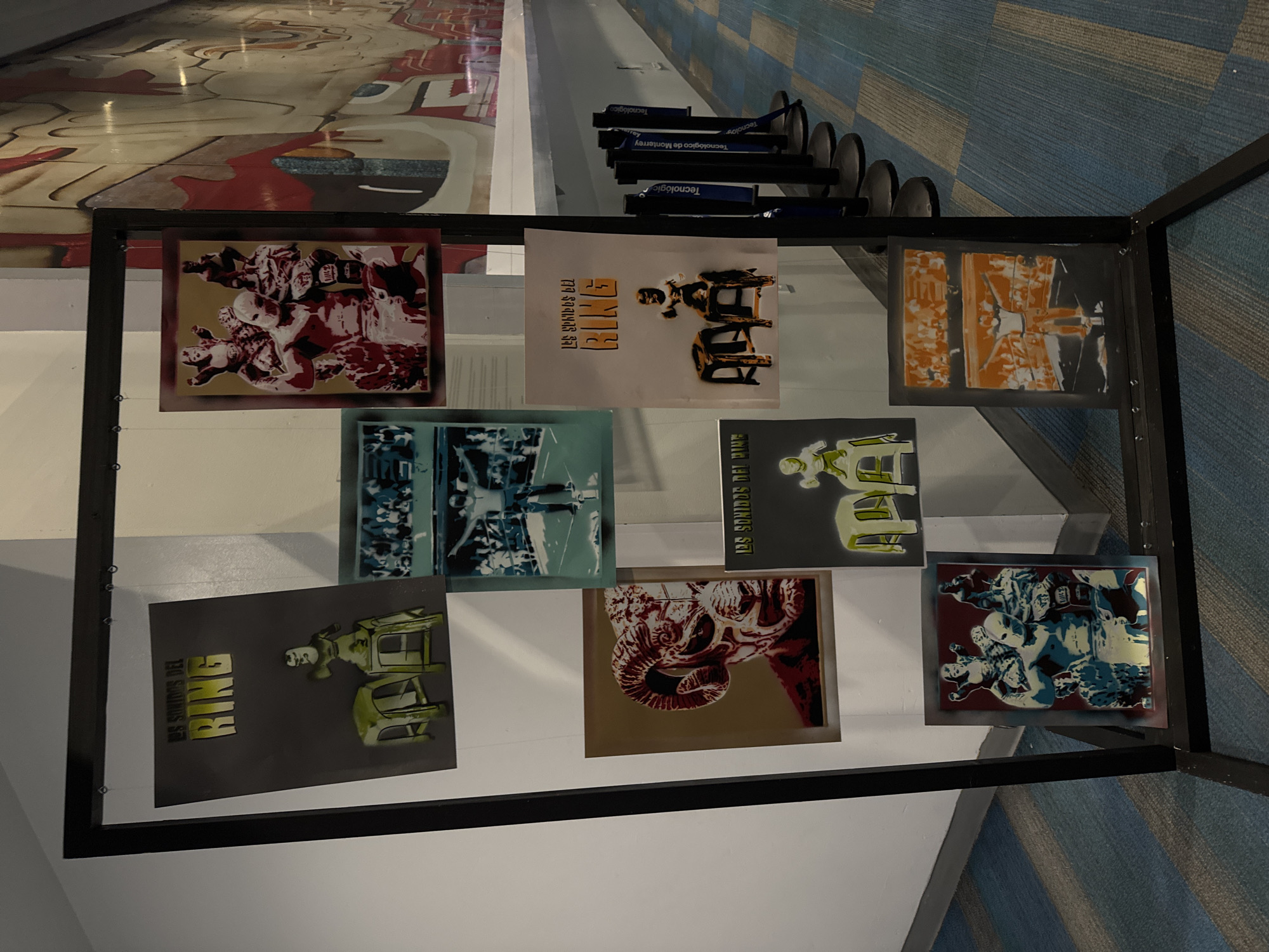 A collection of nine posters in a black metal frame is displayed indoors, likely an exhibition. The posters feature stylized graphic designs related to boxing, with the words "RING," "BIKE," "US SONIDOS DEL RING," and "LES SONIDOS DEL PINE" appearing on some. The color palettes include reds, golds, greens, blues, and blacks. The posters are arranged in a 3x3 grid within the frame, which is set on a white and gray surface. In the background, there is a floor with an abstract design of red, gray, and white shapes. Behind the framed posters, there are objects labeled "Tecnológico de Monterrey," indicating the location may be related to that institution, likely in the city of Puebla, Mexico. The floor transitions to a blue and gray patterned carpet.