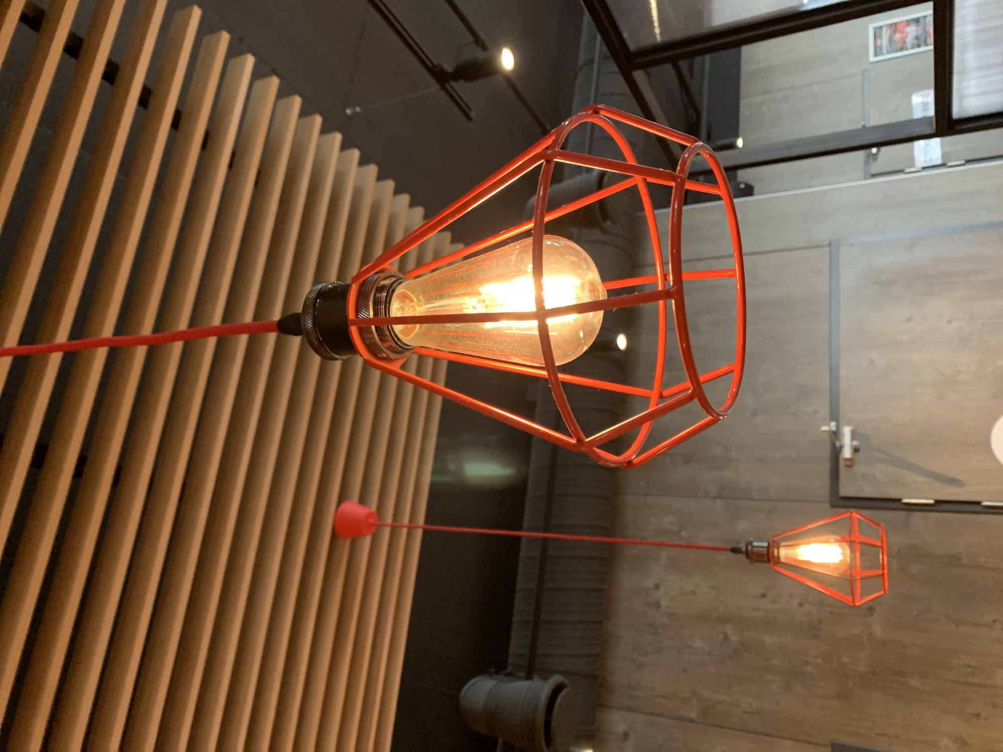 The image shows an indoor scene with two red pendant lamps hanging from a slatted ceiling. The lamps have a modern, industrial design with exposed bulbs enclosed in red geometric cages. The larger lamp is in the foreground and is in sharp focus, while the smaller lamp is in the background and slightly out of focus. The ceiling is made of light-colored wooden slats, and the walls appear to be a textured gray material, possibly concrete or stone. In the background, there is a closed door with a metal handle, and a glimpse of a framed picture on a wall. The lighting in the scene is warm and inviting, coming primarily from the lamps themselves. The overall impression is one of a contemporary interior space, possibly a restaurant or cafe. There are no people visible in the image. There is no information available to determine the location in the city of Pinsk, Belarus.