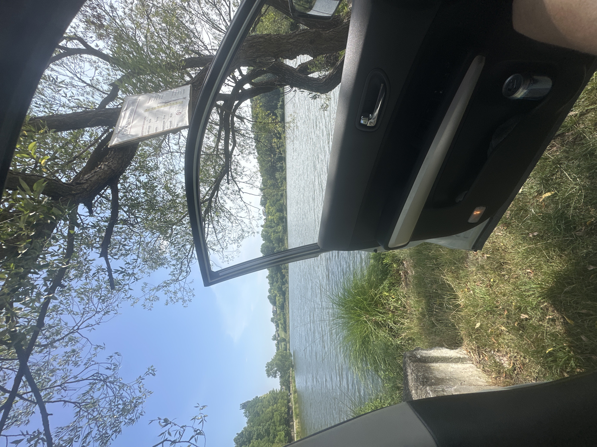 This image depicts a view from inside an open car door, looking out towards a body of water. The car is parked on a grassy area next to the water, with trees and foliage surrounding the scene. A sign is attached to a tree in the middle ground. The sky is clear and blue with some clouds. The scene appears to be a natural environment, possibly a park or a rural area, and based on the provided context, it is located in Makiv, Ukraine. The water is rippling, suggesting a gentle breeze. The car door is open, revealing the interior of the car, including a door handle and a storage compartment with a can inside. The overall impression is one of a peaceful, outdoor setting, perhaps a recreational spot.