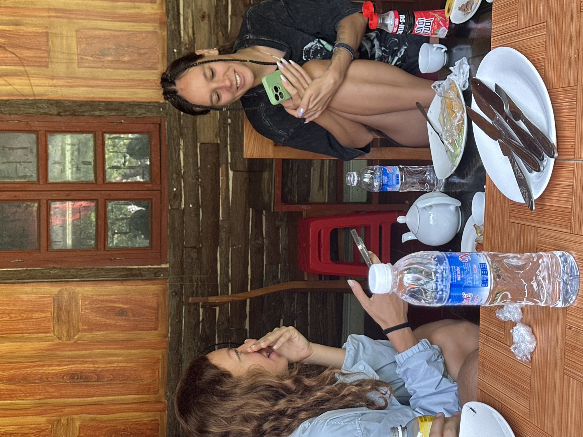 Two women are at a wooden table inside what appears to be a rustic establishment in Mèo Vạc Commune, Vietnam. One woman, wearing a dark t-shirt and braids, is sitting with legs crossed, holding a green smartphone, and smiling. The other woman, with curly brown hair, is wearing a light blue shirt and holds a phone or bottle, her hand near her face. On the table are plates, cutlery, water bottles, a teapot, condiments, and food remnants. The room features wooden walls and a window showing greenery outside.