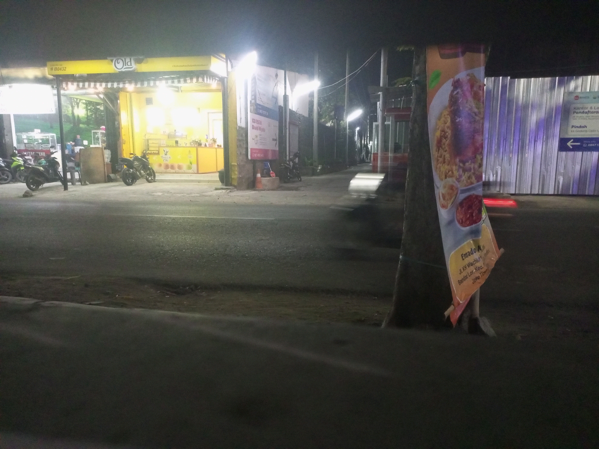 The image shows a nighttime scene on a street in Kediri City, Indonesia, likely near a commercial area. In the foreground, a tree with a banner on it stands on the right side of the frame. The banner displays an image of food and text in Indonesian, reading "Emado Ka" and "Jl. KH Wachid Hasyim, Bandar Lor, Kec. Mojoroto, Jawa Timur." To the left of the tree, a brightly lit yellow food stall with the "Old" logo is visible, featuring a counter, display cases, and outdoor seating. Several motorcycles are parked in front of the stall. Further down the street, there are buildings and streetlights. A blurred vehicle appears to be moving on the road. The overall environment is dimly lit, typical of a street at night, with artificial lighting from the stall and streetlights illuminating the scene.