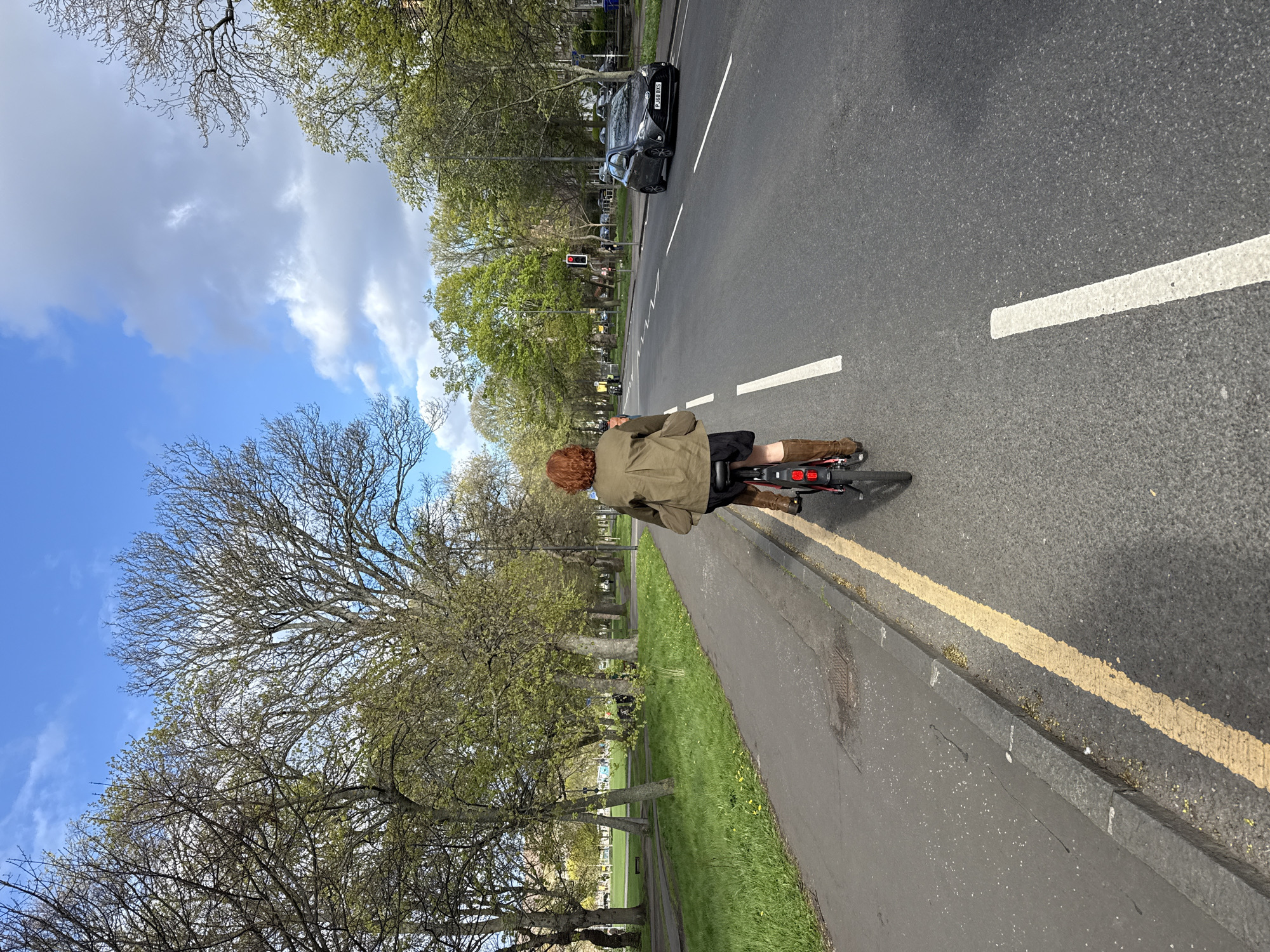 A person is riding a bicycle along a street in an urban environment. The individual is wearing a khaki jacket, dark shorts, and brown boots. They have red hair. The bicycle has a red rear light. The street has yellow lines indicating lane markings. To the left of the bicycle rider is a grassy area with trees, and further left is a tree line with a building in the distance. On the road to the right, a black car is visible further down the street. There is a traffic light ahead. The sky is visible above the trees, with a mix of blue sky and clouds. The image appears to be taken in a city environment, location unknown.