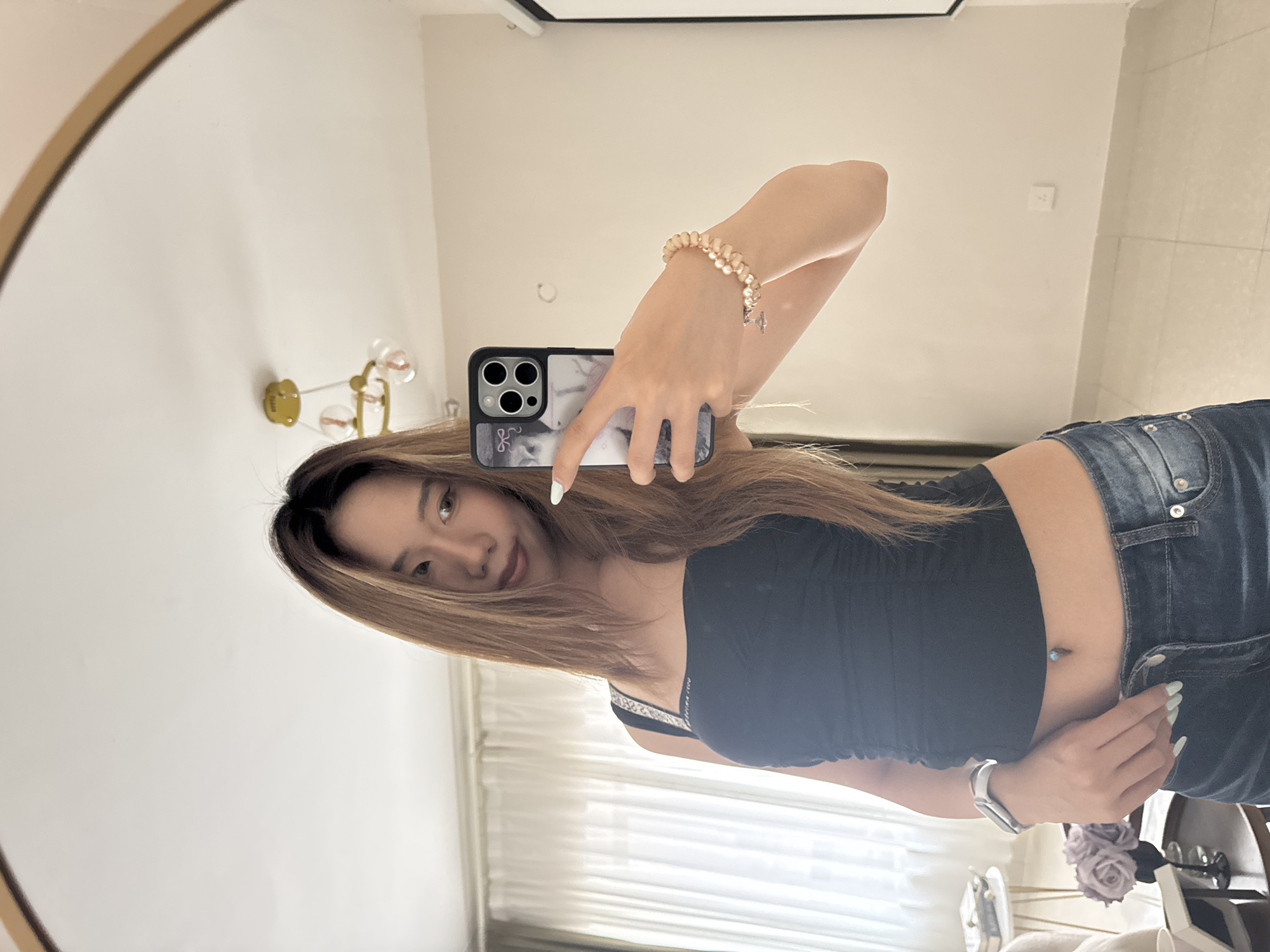A young Asian woman is taking a mirror selfie in what appears to be a bedroom. She has long, light brown hair and is wearing a black ruched tank top, blue jeans, and a silver watch. She is also wearing a pearl bracelet on her right wrist and has a belly button piercing. In her right hand, she holds a smartphone with a marble-patterned case. The background shows a large mirror, sheer curtains, a light fixture, and some furniture including a small table with books and flowers. The floor is tiled and the walls are painted a neutral color. The image is well-lit, suggesting it was taken during the day.