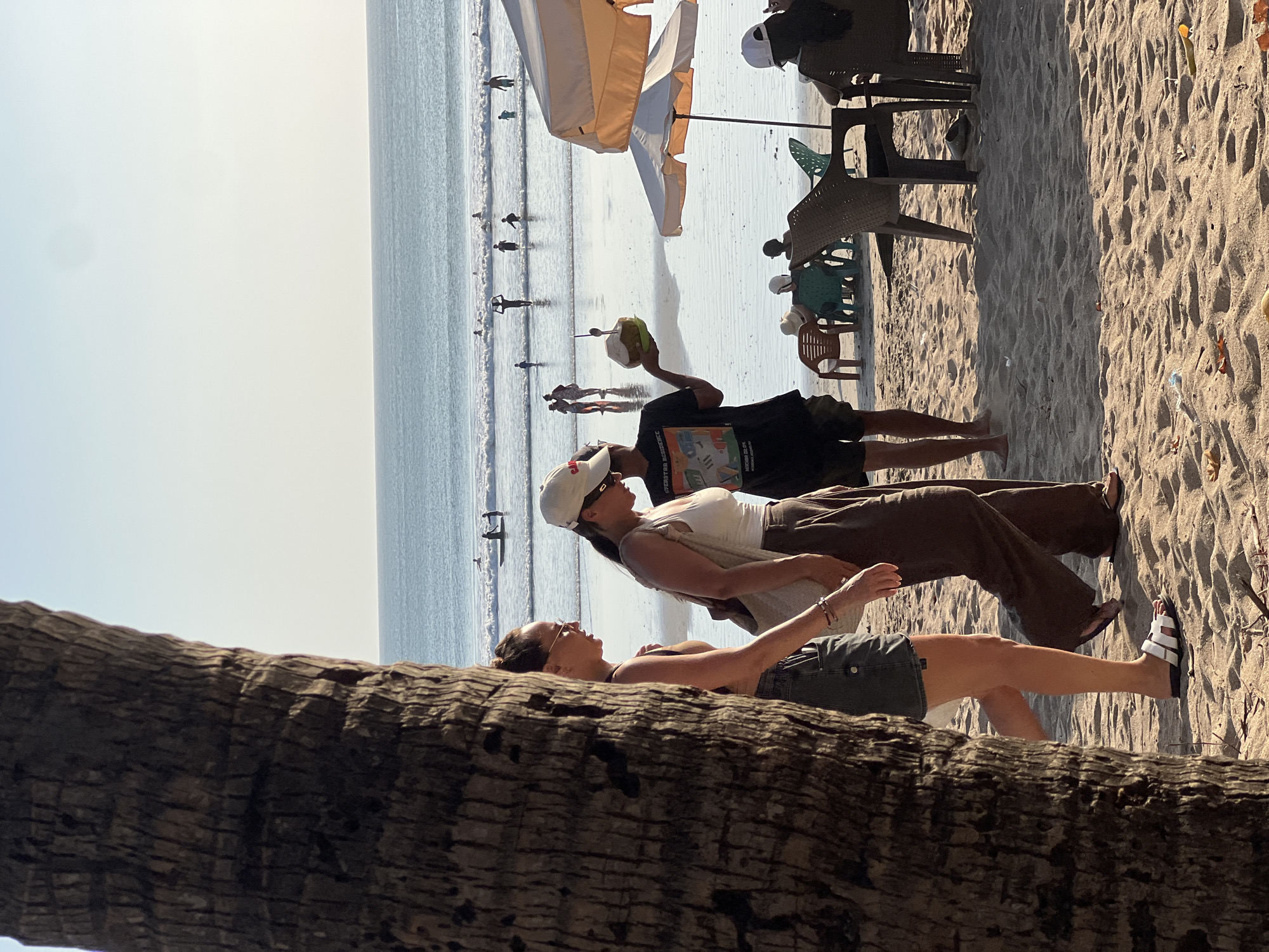 The image depicts a sunny beach scene in Kuta, Indonesia. In the foreground, the textured bark of a large palm tree is visible on the left side. Two women are walking from left to right across the sandy beach. The woman on the left wears sunglasses, a dark top, and denim shorts. The woman on the right wears a white cap, a white tank top, and brown pants. Behind them, a man holds up a coconut drink with a straw. Further in the background, the ocean stretches to the horizon, with several people swimming and standing in the shallow water. Beach chairs and umbrellas are set up on the sand, indicating a popular tourist spot. The overall atmosphere is relaxed and typical of a beach day.
