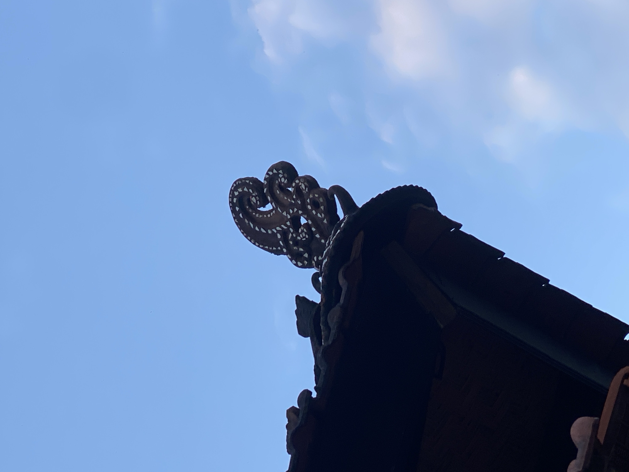 An ornate, brown, octopus-shaped finial adorns the roof of a building against a bright blue sky with wispy white clouds. The finial has intricate details, with white markings suggesting eyes or suckers. The roof itself is tiled and appears dark brown, with a portion of a wooden eave visible beneath it. The image was taken in Kuta Selatan, Indonesia, as suggested by the architectural style and the presence of a Hindu temple element.