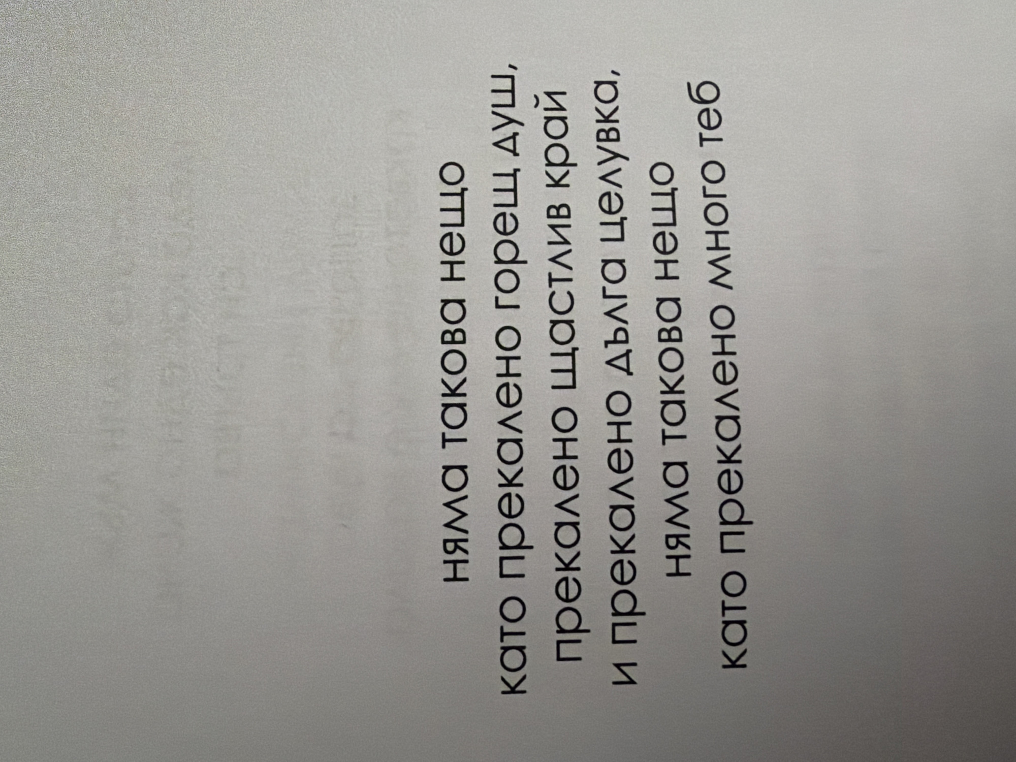 The image shows a page from a book with text printed in Bulgarian. The text is in a dark font on a light-colored, slightly textured paper. The text is centered on the page and appears to be a quote. The quote reads: "няма такова нещо като прекалено горещ душ, прекалено щастлив край и прекалено дълга целувка, няма такова нещо като прекалено много теб". This translates to: "There is no such thing as too hot a shower, too happy an ending, and too long a kiss, there is no such thing as too much of you." The background of the page is slightly out of focus, but appears to be a plain, perhaps slightly reflective surface. There are no people, objects, or environmental elements visible other than the book page. The image does not provide any information about the location in the city of Pazardzhik, Bulgaria.