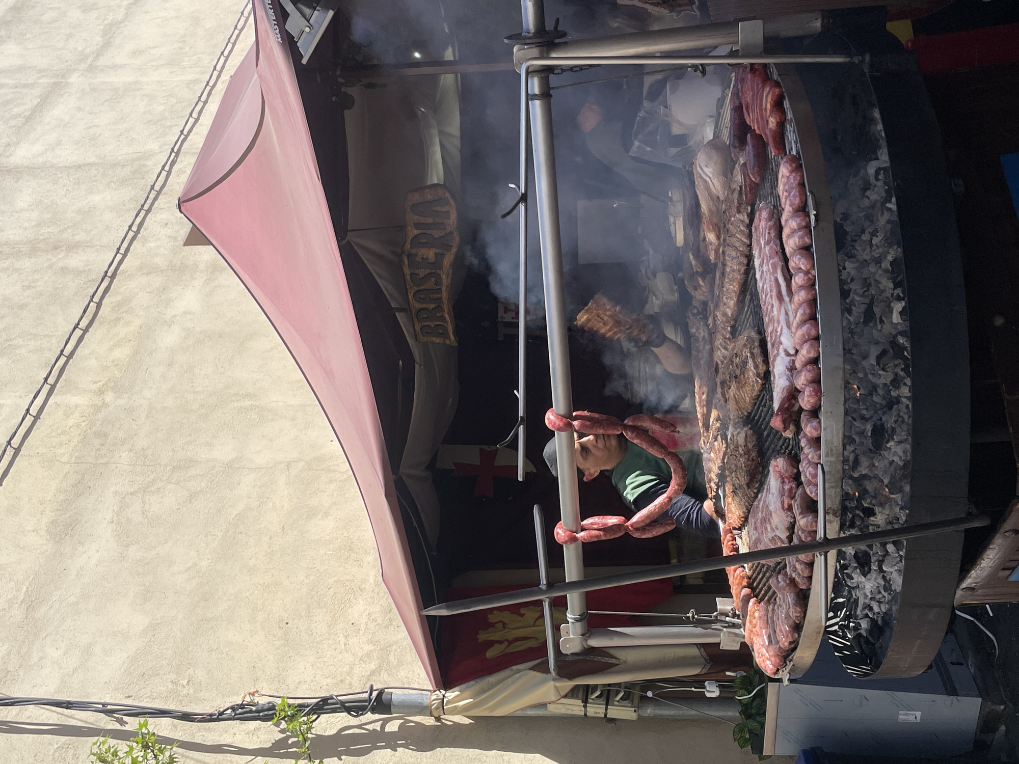 The image captures a street food stall in Hostalric, Spain. A large, round grill filled with various meats, including sausages and ribs, dominates the scene. Smoke rises from the grill. A person in a green shirt is attending the grill. A red awning covers part of the stall. A sign reads "BRASSERIA." A flag is visible under the awning. The stall is set against a light-colored building wall. Power lines and some foliage are visible at the bottom of the image.