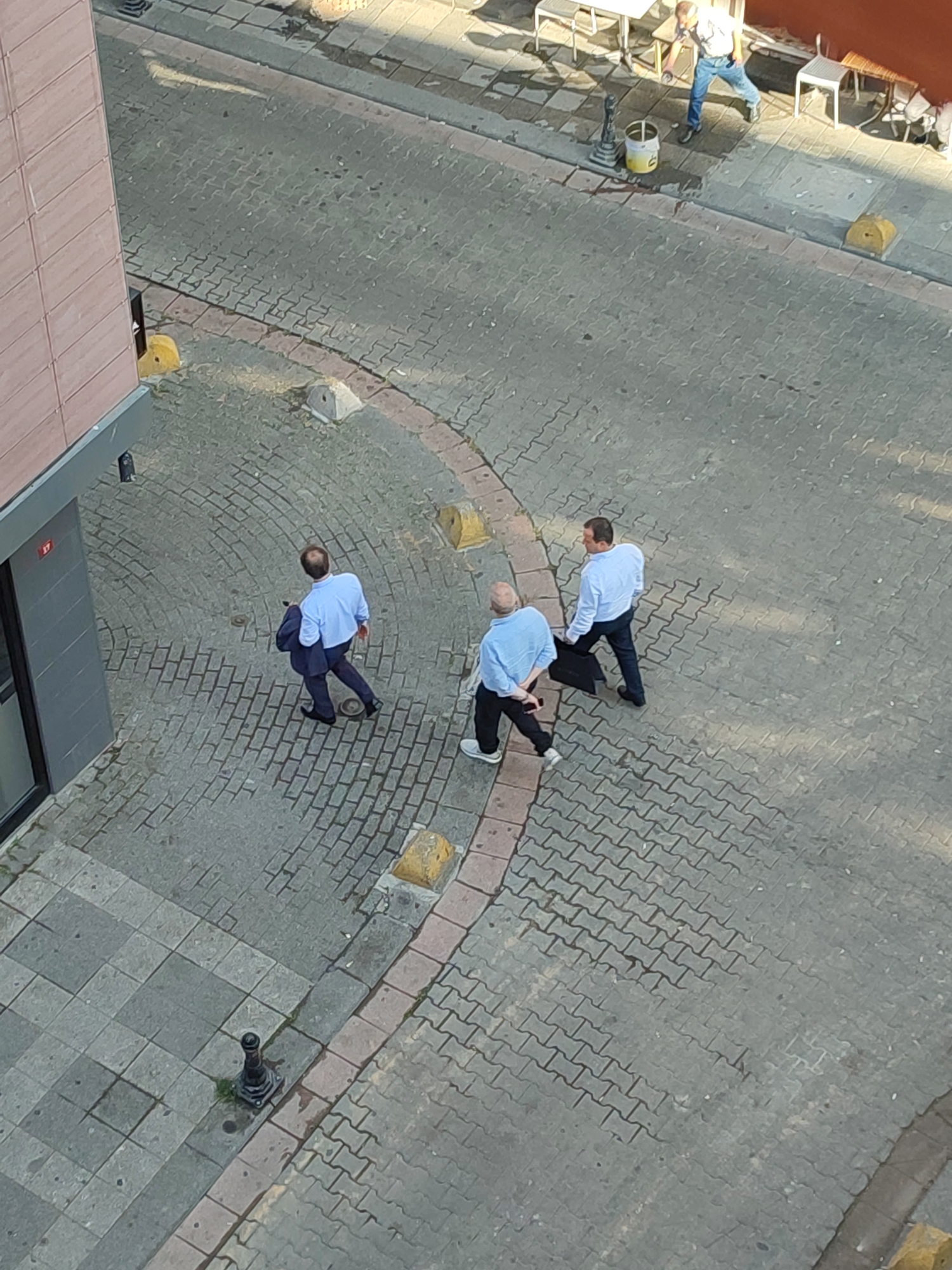 Three men in light blue shirts and dark pants walk on a cobblestone street in Kadıköy, Turkey. The man on the left carries a dark jacket over his left arm and walks away from the camera, looking down. The middle man walks towards the camera, carrying a dark briefcase in his right hand and looking forward. The man on the right also walks towards the camera, looking forward. A building with a pink facade is visible on the left edge of the image. Yellow concrete bollards are placed along the edge of the street. In the background, two white tables and chairs suggest an outdoor seating area.