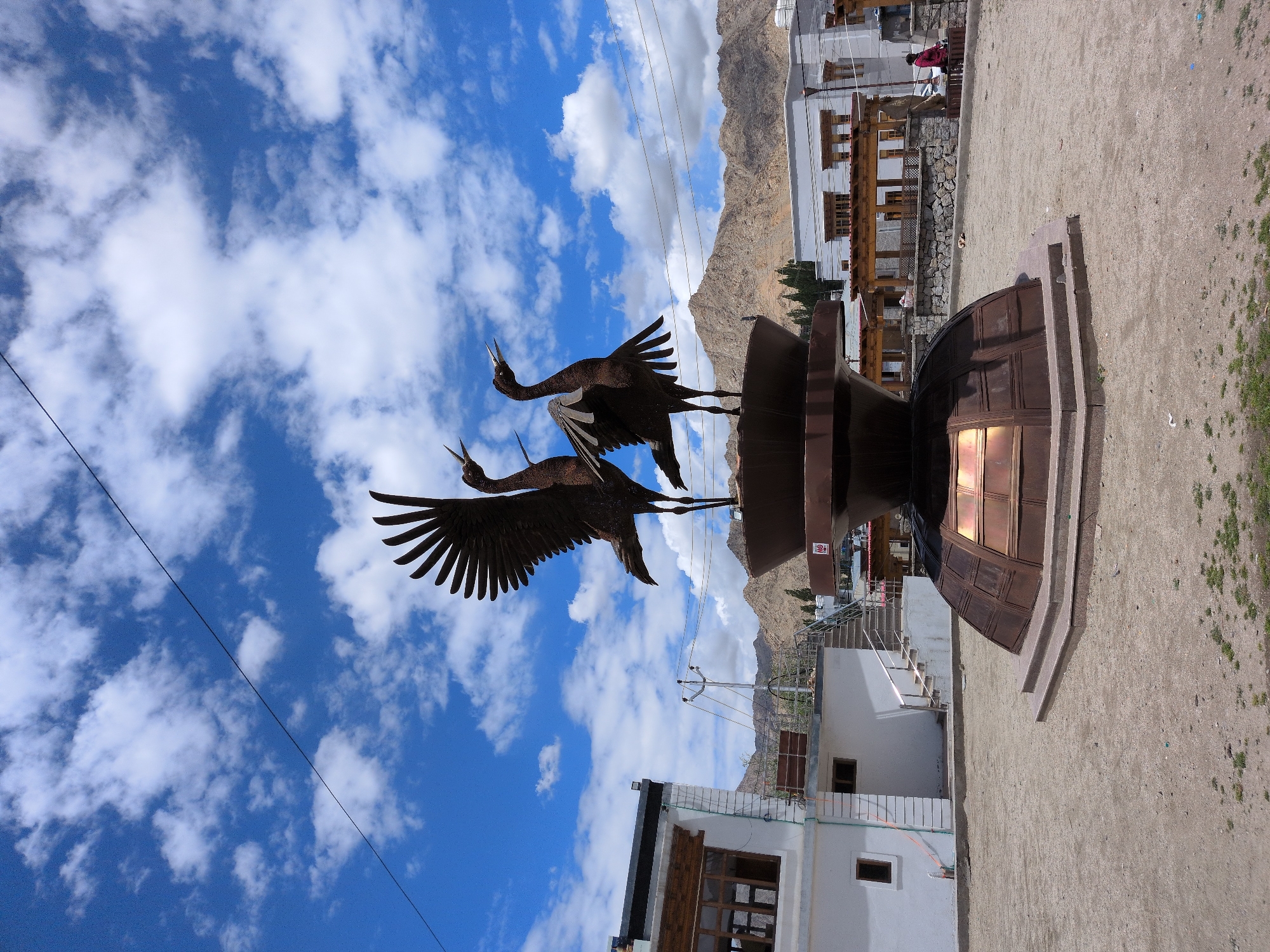 The image shows a large metal sculpture of two cranes with their wings outstretched, positioned in the center of an outdoor paved area. The sculpture is mounted on a tiered, octagonal base made of a darker metal, possibly copper or bronze. Behind the sculpture, there are several buildings with white and light-colored facades, featuring wooden window frames and balconies. In the background, a range of barren, rocky mountains under a bright blue sky with scattered white clouds is visible. A single black power line stretches diagonally across the upper portion of the sky. The paved area appears to be a public space, with some sparse vegetation and loose gravel on the ground. In the right side of the image, a woman in a red jacket is walking away from the camera. The scene is consistent with the public spaces found in the city of Leh, Ladakh, India, known for its mountainous terrain and distinct architecture.