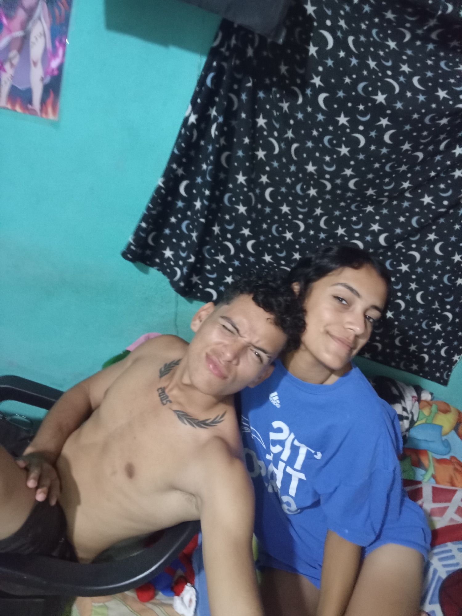 Two individuals, a male and a female, are indoors. The male is shirtless, seated in a black chair, and has a tattoo on his upper chest. He is grimacing at the camera. The female is wearing a blue t-shirt and is smiling. They are close together, potentially touching. The background includes a teal wall, a poster, a black curtain with a star and moon pattern, and a colorful blanket. The specific location in Altagracia, Venezuela, is not identifiable from the image.