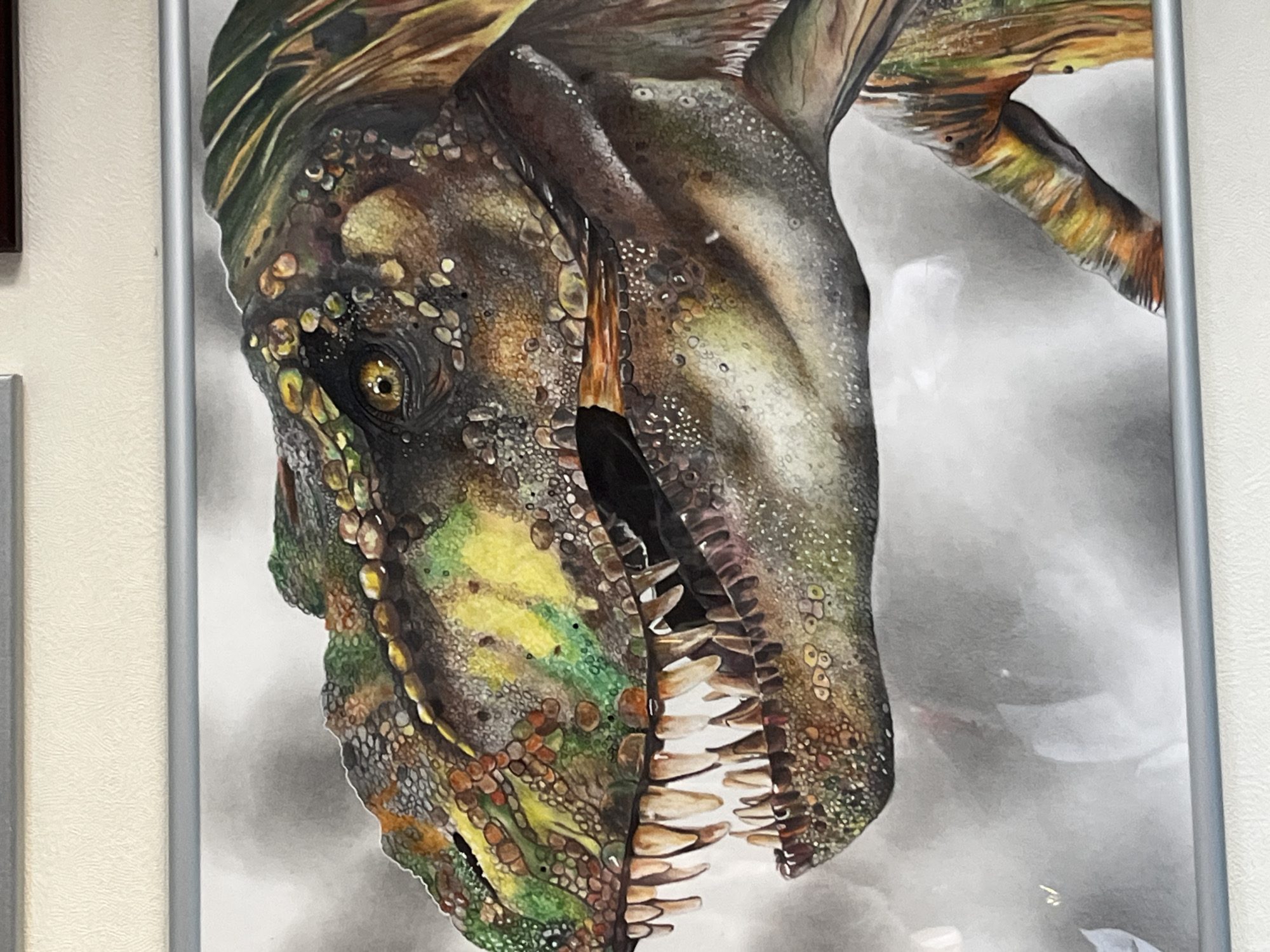 A framed picture of a dinosaur's head is hanging on a wall. The dinosaur is depicted with a variety of colors and textures, including green, brown, yellow, and black, suggesting scales or skin. Its mouth is open, revealing sharp teeth. The background of the picture is a muted grey. The location is not provided.