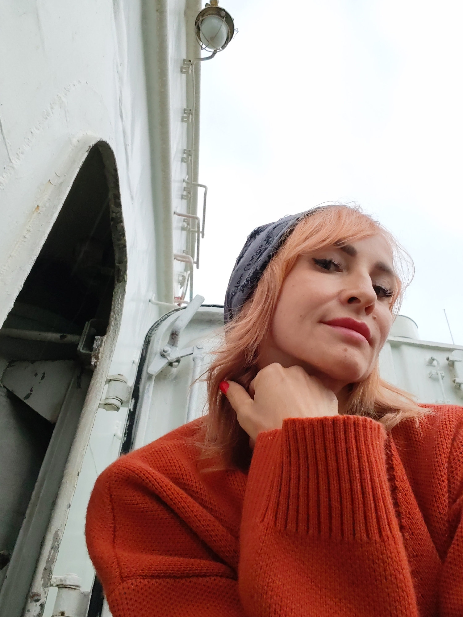 A woman with reddish-blonde hair, wearing a grey bandana and an orange sweater, is taking a selfie. She has her left hand on her chin, and her right hand is partially visible. The background appears to be the exterior of a large, white, industrial structure, possibly a ship or a building with an industrial aesthetic. There's a dark opening to the left of the woman, and some metallic fixtures and a light fixture are visible on the structure. The sky is overcast. The location is not specified to be in Aleksandrovskoe, Russia.