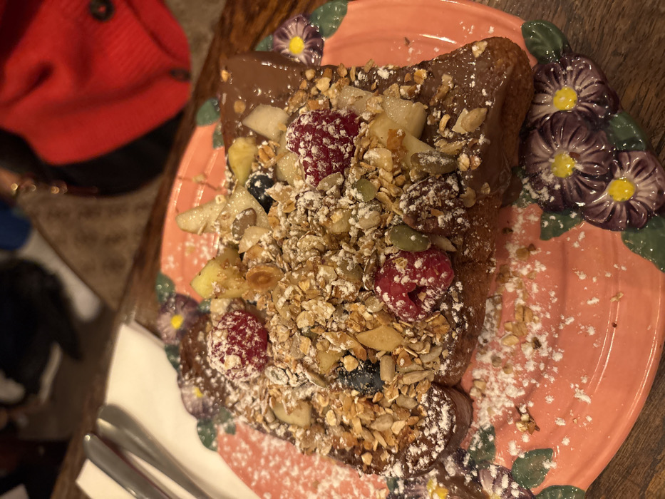 A rectangular slice of French toast, heavily garnished, is presented on a pink ceramic plate. The toast is covered with a melted dark brown spread, likely chocolate or hazelnut, over which a generous layer of granola is scattered. The granola consists of rolled oats, various seeds (including pumpkin and sunflower), and chopped nuts. Integrated into the granola and spread are whole red raspberries, dark blueberries, and diced light-colored fruit pieces, appearing to be pear and apple. A dusting of white powdered sugar is distributed across the entire dish and lightly on the plate's rim.
The ceramic plate features a raised, textured rim adorned with purple and yellow floral designs, specifically three flowers visible on the left and two on the right. In the upper left quadrant of the image, silverware consisting of a fork and knife rests on a white napkin. The immediate environment suggests a table made of dark wood, visible as a blurred surface.
In the blurred background, several figures or objects are partially visible. To the upper right, a person is seated, identifiable by a vibrant red garment, possibly a sweater or jacket. To the upper left, another indistinct, blurred figure or object shows a white element, possibly part of footwear or clothing. The overall setting is indoors, likely a cafe or restaurant, characterized by dim lighting. The exact location within Paris, France, is not discernable from the visual information presented in the image.