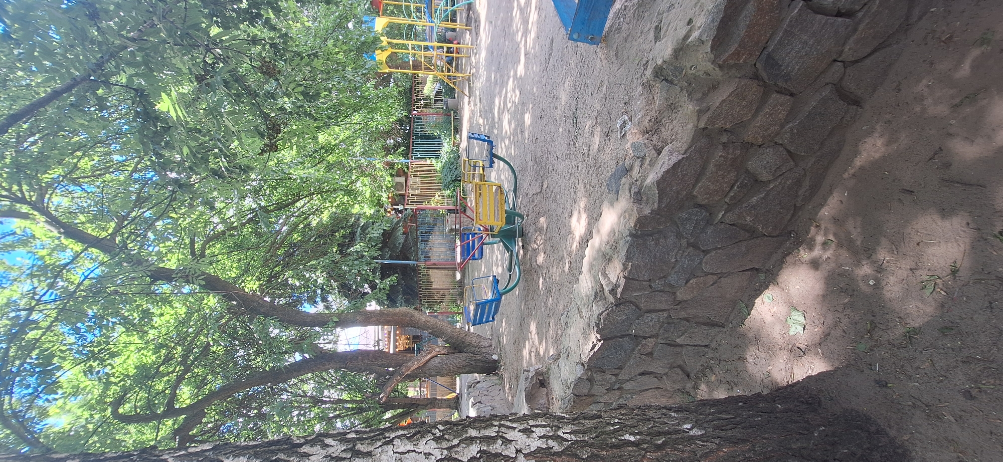 The image shows a playground scene on a sunny day, likely in Bila Tserkva, Ukraine. A vibrant yellow and blue merry-go-round with four seats is the central object, positioned on a dirt ground. Behind it, a colorful playground structure with yellow and red elements is partially visible, along with a metal fence. Lush green trees with a visible trunk of a large tree in the foreground, covered in rough bark, dominate the left side of the image, providing shade. The ground is covered in dirt, with scattered leaves and some debris. A low stone wall lines the right side of the playground area. The sunlight casts dappled shadows through the trees onto the ground. There are no people visible in the frame. The overall impression is a peaceful, sunlit outdoor recreational space.
