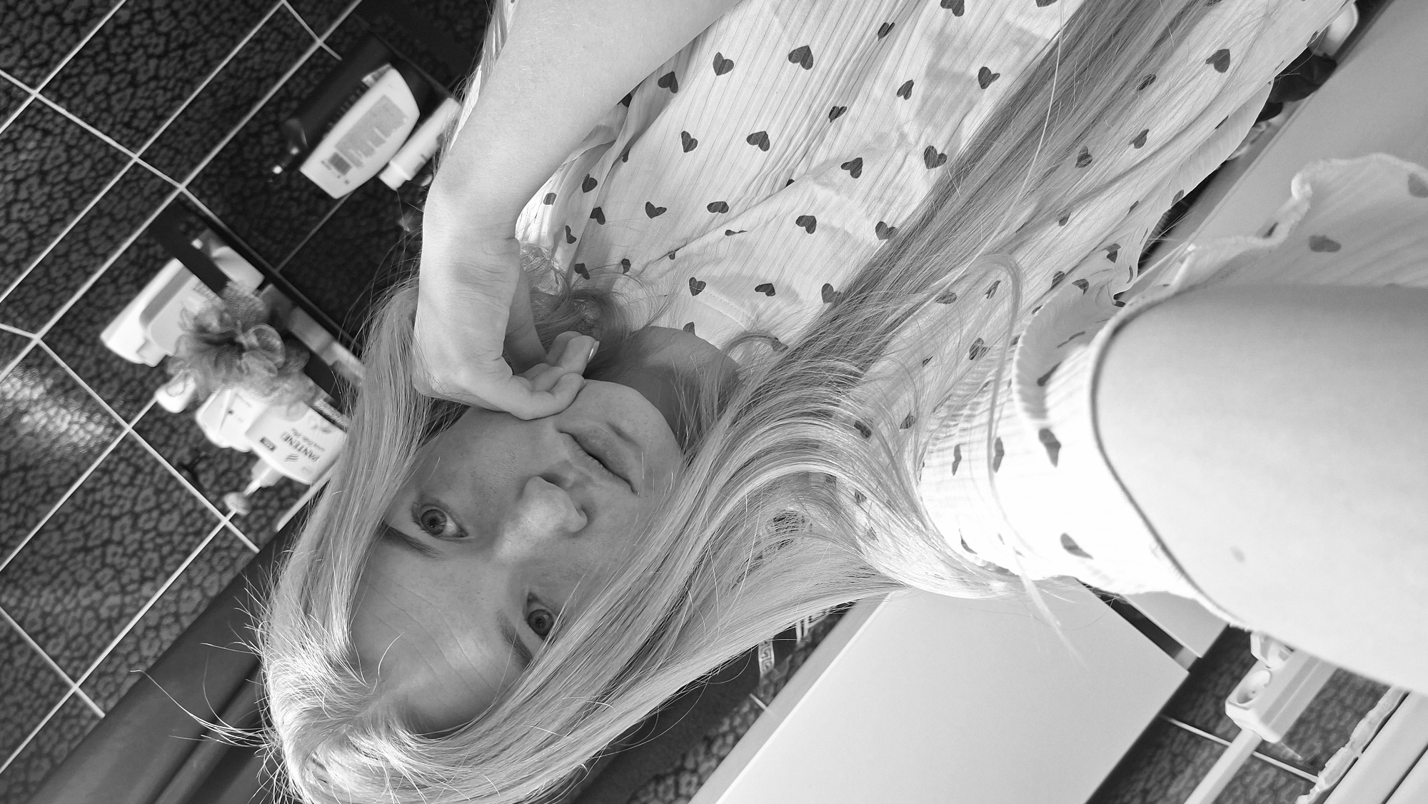 A black and white, skewed perspective, selfie-style photograph captures a fair-skinned person with long, light-colored hair. The person is resting their face on their hand. They appear to be in a bathroom setting.
Details include:
- The person is wearing a light-colored garment patterned with dark hearts.
- The background features a tiled wall with a grid pattern, and a shelf-like structure with various toiletries and a shower loofah.
Location: Based on visual evidence, including the tiled bathroom setting and toiletries, the photograph was taken indoors in a bathroom. However, no information indicates the image was taken in the city of Chaikovsky, Russia. It is based on image features alone.