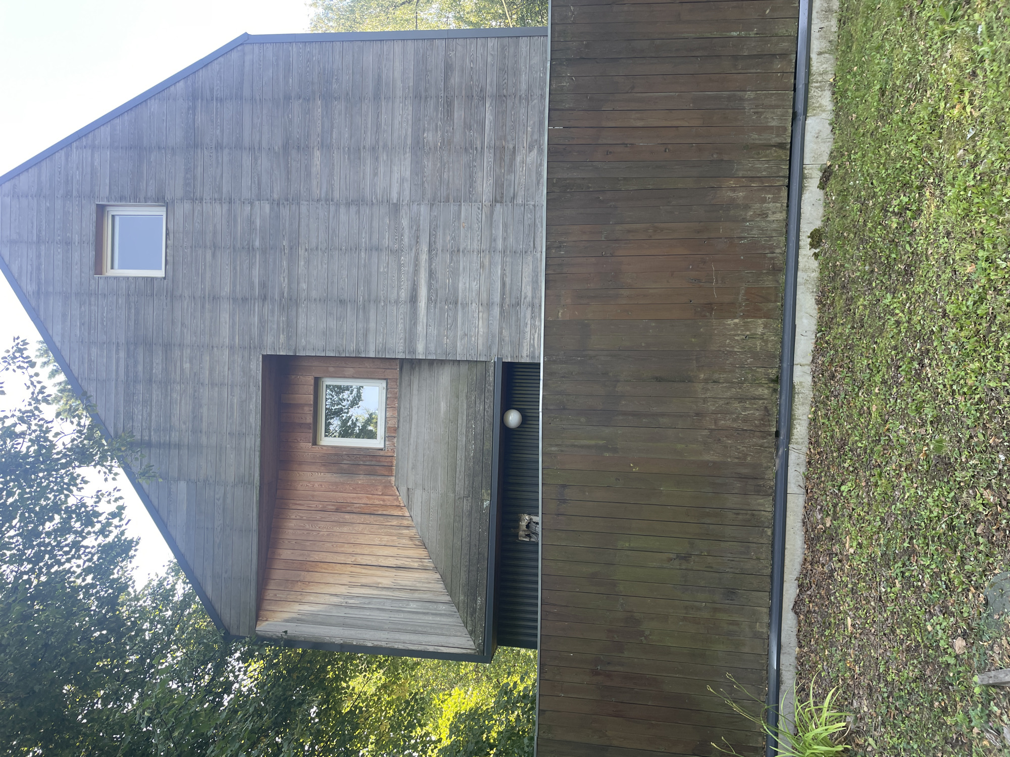 The image depicts the exterior of a modern wooden house, likely in Vidnoye, Russia, as indicated by the context. The house features a unique architectural design with contrasting wooden cladding. The upper portion of the facade is clad in weathered, gray horizontal planks, while a prominent angled section is covered in warm-toned vertical wooden planks. Two square windows are visible on the facade. One is positioned high up on the gray section, and another is located within the wooden angled section. A dark wooden fence runs horizontally across the foreground, partially obscuring the lower part of the house. Beyond the fence, a patch of overgrown grass and fallen leaves covers the ground. Trees with green foliage are visible to the left and right of the house, suggesting a natural or suburban setting. The sky is visible at the top of the image and appears to be clear. There are no people or animals in the frame.