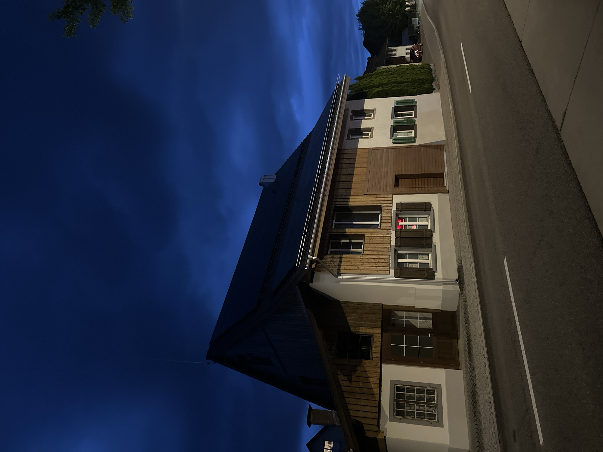 The image captures a quiet street scene in Embrach, Switzerland, during deep twilight or night. A multi-story house is prominently featured on the left, oriented at an angle, revealing its facade and roofline.
The house displays traditional and modern elements. Its steep, dark-tiled roof is equipped with what appear to be solar panels or lightning protection rods running along the ridge. Sections of the building's exterior are clad in warm brown wooden siding, while other parts are painted a light, possibly off-white, color. Numerous windows are visible across the facade; many have traditional wooden shutters, some open, some closed, in shades of green and dark brown. A chimney stack is visible on a lower roof section towards the bottom left. Faint internal lighting can be seen through some windows, and a reddish glow, possibly from a sign or decoration, is visible through a ground-floor window on the right side of the house.
To the right of the house is a smooth, dark asphalt road with a distinct white dashed line running down its center. A patterned grey curb or paved area separates the road from the house. In the background, across the street, other buildings are partially visible behind a well-maintained green hedge and trees. A small, dark-red vehicle can be faintly seen parked further down the street.
The sky is a deep, rich indigo blue, characteristic of dusk or night, with subtle cloud formations. Dark green foliage from trees can be seen in the upper left corner and behind the house in the distance. The scene is devoid of people or direct activity, conveying a calm and tranquil atmosphere. No discernible text is visible.