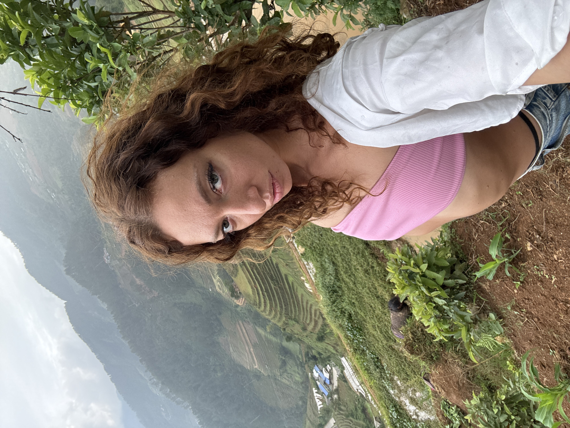 A young woman with long, curly brown hair and fair skin is posing for a photograph in the city of Dế Xu Phình, Vietnam. She is wearing a pink top, denim shorts, and a white shirt. She is standing on a hillside overlooking rice terraces and mountains, surrounded by greenery, including trees and vegetation. The sky is overcast.