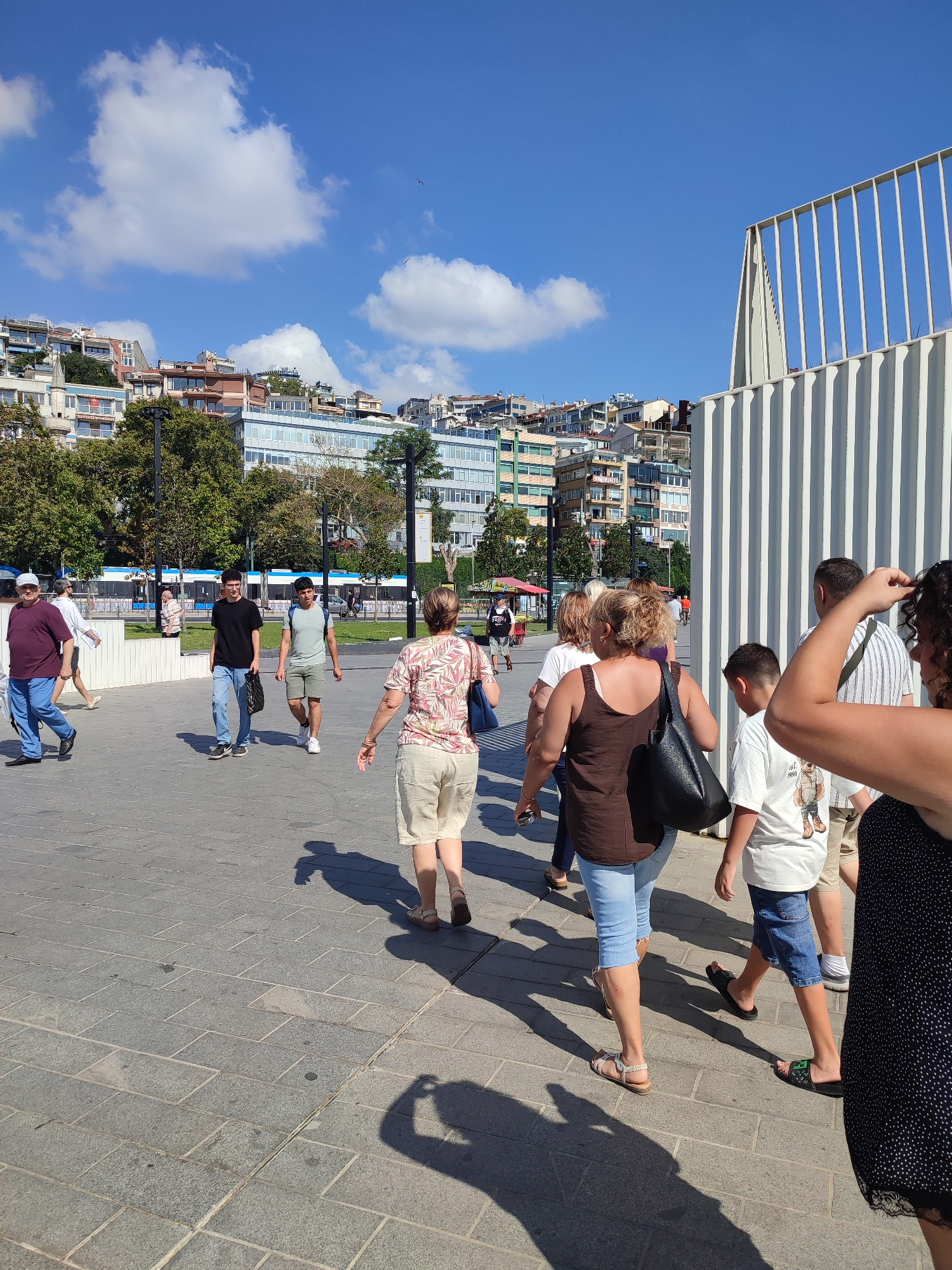 A bright, sunny day in Beyoğlu, Turkey, shows a group of people walking on a paved plaza with buildings and trees in the background. In the foreground, a woman with curly hair is partially visible on the right, wearing a black and white polka dot dress. To her left, a young boy in a white t-shirt and denim shorts walks away from the camera, and next to him, a woman in a brown tank top and blue capri pants carries a black purse. Behind them, a man in a striped shirt walks towards the right. Further back, two young men walk towards the viewer; one in a black t-shirt and jeans, and the other in a light green t-shirt and khaki shorts. An older man in a maroon t-shirt and blue pants walks away from the camera on the left. A woman in a floral print shirt and beige shorts is also seen walking away from the camera. In the mid-ground, trees line a grassy area with a blue and white tram passing by. The background features a densely packed cityscape with multi-story buildings under a clear blue sky with scattered white clouds. A modern white structure with vertical slats is visible on the right side of the image.