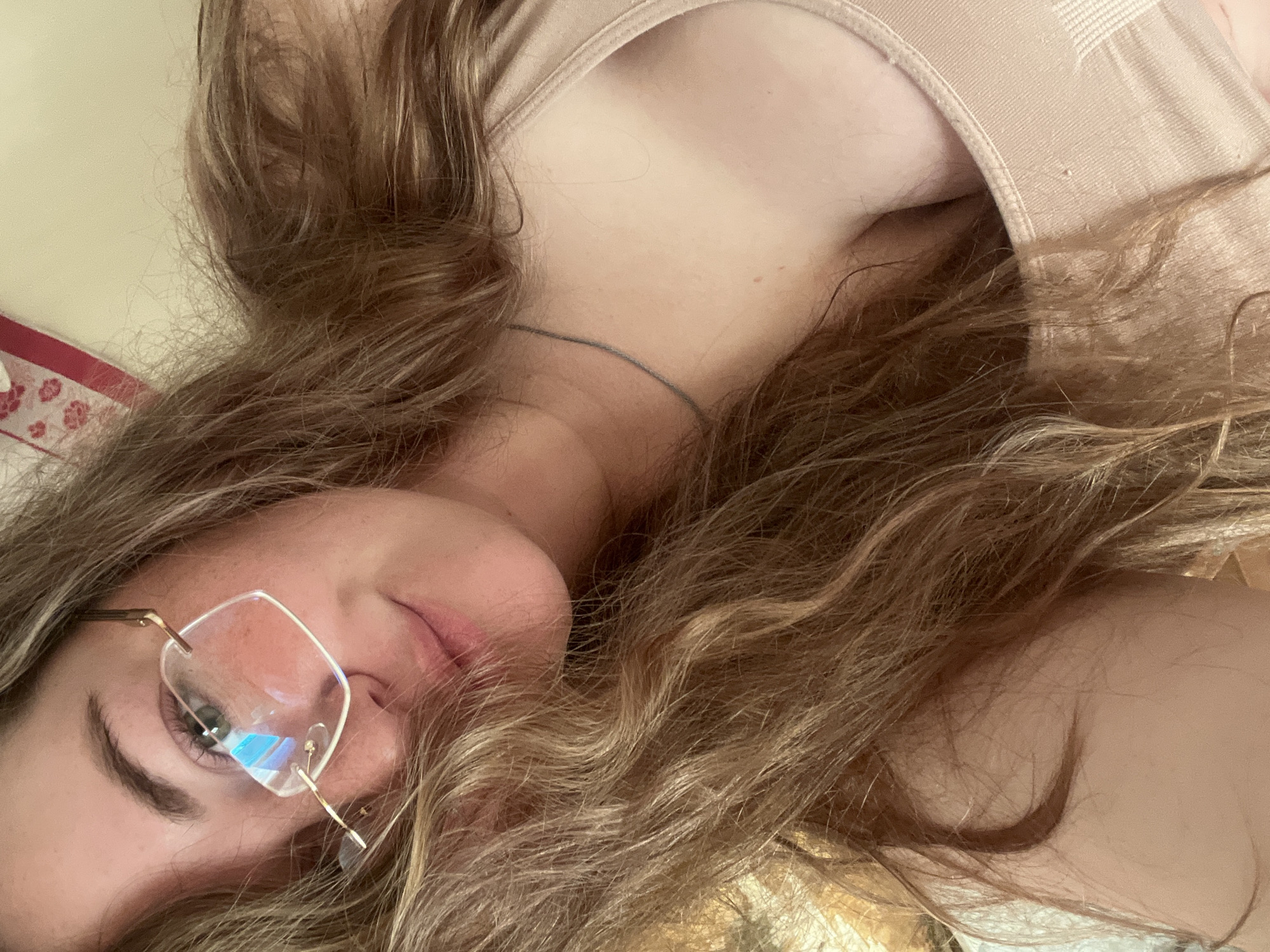 A woman with wavy, light brown hair is lying on her back, facing the camera at an angle. She is wearing rimless glasses with gold frames. She has a necklace and is wearing a light-colored top. The background includes what appears to be a floral patterned object. There is no indication of being in Сосново, Russia.
