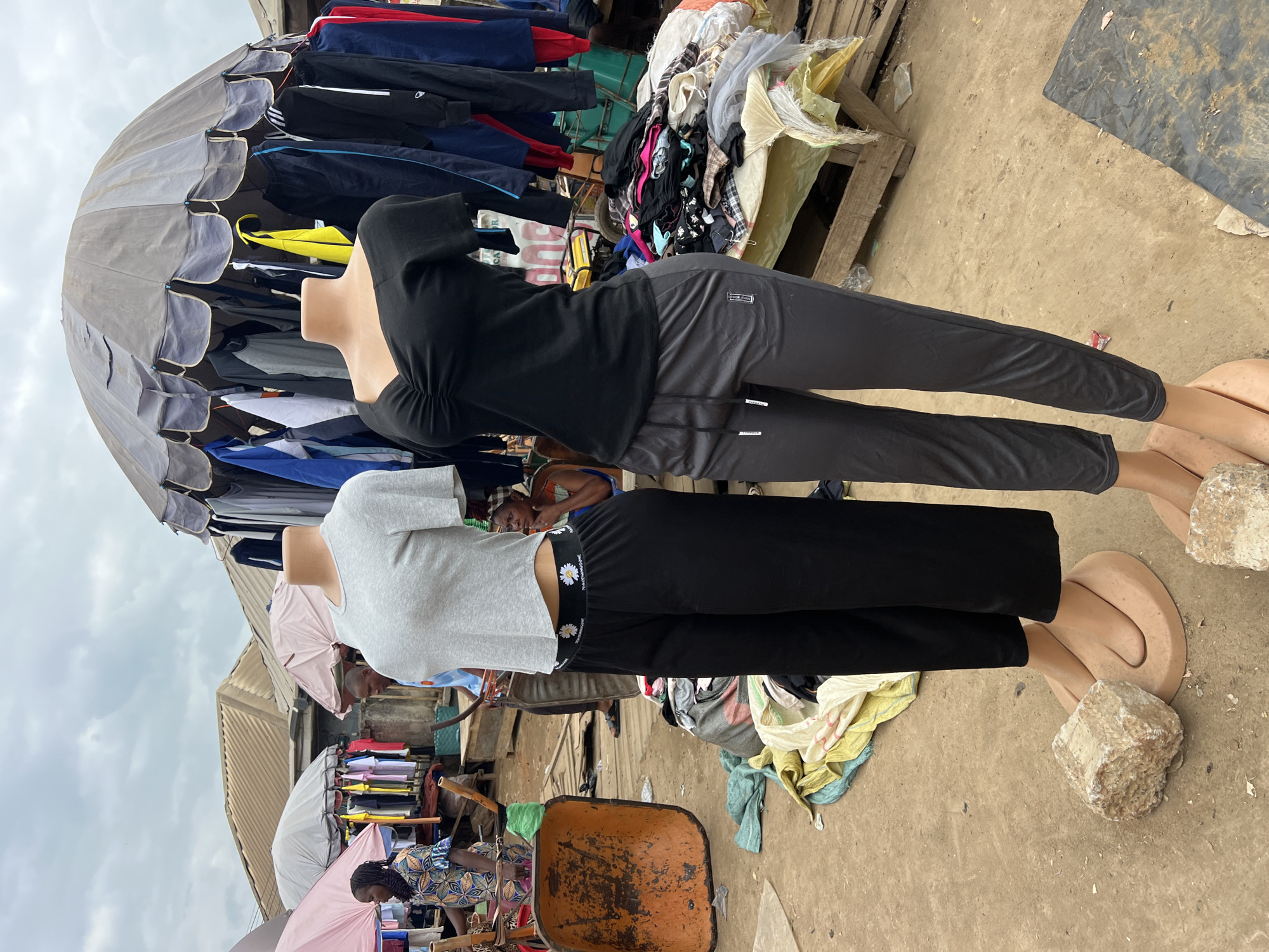 The image depicts an outdoor market scene in Asaba, Nigeria. The primary subjects are two mannequins, one in a black hooded top and dark gray pants, and another in a light gray t-shirt and black pants with a daisy pattern waistband. The mannequins are positioned vertically and are part of a clothing stall.
Behind the mannequins, a variety of clothing items are displayed on hangers under a large, grey umbrella. The clothing appears to be casual wear, including t-shirts, jackets, and pants. To the right of the stall, there are wooden pallets with more piles of clothing and fabric. In the background, other market stalls and structures are visible, suggesting a busy commercial area. A wheelbarrow is prominently featured in the lower right portion of the image.
The ground is unpaved and dusty, typical of an outdoor market. The sky is overcast, indicating cloudy weather. The overall impression is that of a typical day in a bustling Nigerian market, with vendors selling clothes and shoppers likely browsing. There is no specific activity or event taking place beyond the general operation of the market.
Some visible text is present on the pants of the mannequin on the right, near the waistband: "POEMINE." The time of day is not clearly discernible, but the lighting suggests it is daytime.