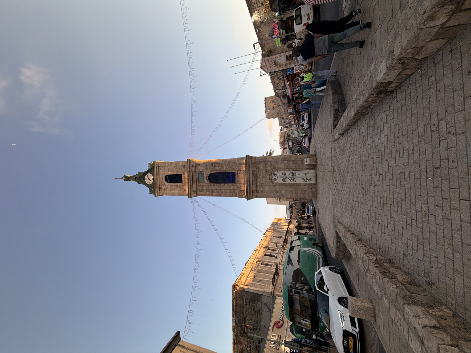 The image depicts the Jaffa Clock Tower located in Jaffa, Tel Aviv, Israel. The clock tower, constructed of light-colored stone, stands tall against a clear blue sky. Suspended on either side are strings of lights. To the right of the tower is a stone-paved street with pedestrians walking and parked cars. A green bus is also parked along the street. Buildings with shops and restaurants are seen along the street.