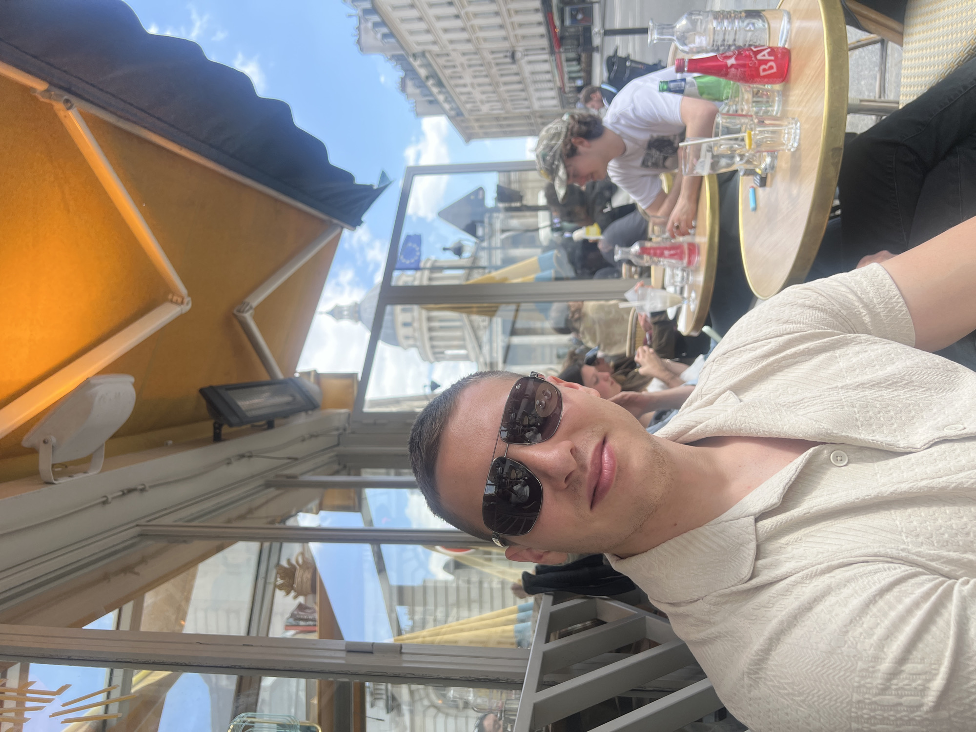 The image captures a vibrant daytime scene at an outdoor cafe in Paris, France. The main subject, a young man, is seated in the foreground, looking at the camera with a slight smile. He wears dark sunglasses that reflect the sky and surrounding buildings, and a light-colored, patterned short-sleeved collared shirt. He has short hair and light facial stubble.
Behind him, other patrons are visible at adjacent tables, enjoying drinks and conversation. One person in a white t-shirt and a camouflage baseball cap is seated prominently at a nearby table. The tables are round with metallic gold rims, holding various beverages including a clear glass bottle of water and a red soda bottle. The red bottle label clearly shows "BAÏSS".
The cafe is equipped with a large, yellow-orange fabric awning supported by metal structures, and features clear glass partitions that reflect the urban environment. A white light fixture and a black heater are mounted on the cafe's ceiling structure.
In the background, quintessential Parisian architecture is visible, consisting of multi-story stone buildings lining a street. A notable landmark, a large domed building resembling the Pantheon, is discernible in the distance through the glass partitions and against the sky. A European Union flag is also visible, potentially as a reflection or positioned nearby, along with a blue and yellow flag.
The bright daylight and clear sky with some clouds suggest pleasant weather. The overall scene conveys a relaxed and enjoyable atmosphere at a bustling city cafe.