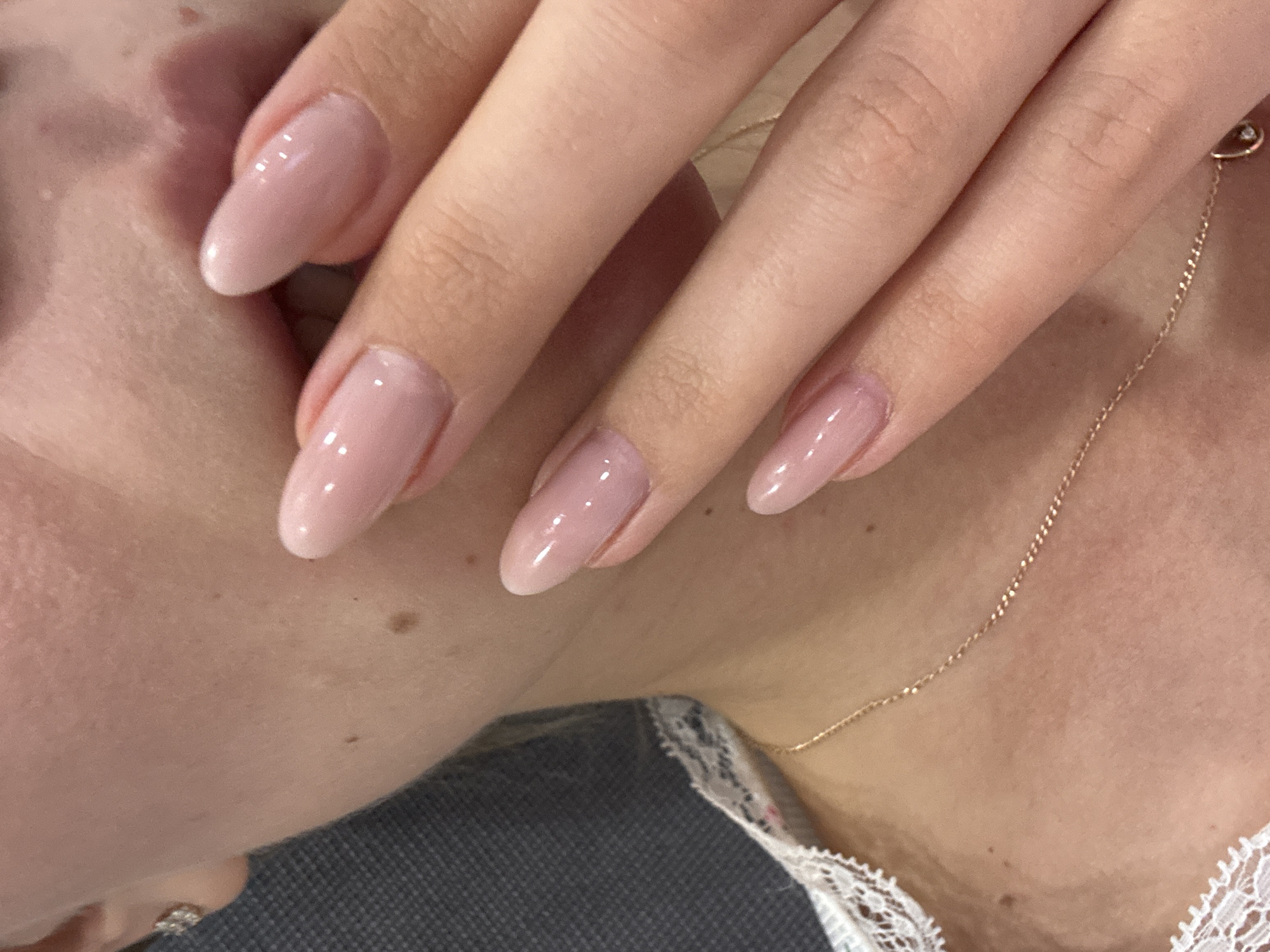 A close-up shot focuses on a person's hand resting near their face and upper torso. The fingernails are elongated and painted with a light pink, glossy nail polish. A thin gold chain necklace adorns the person's neck, with a small, round pendant. The individual is wearing a garment with white lace detailing along the collar. The background is a mix of skin tone and a gray fabric. There is no discernible indication of location.