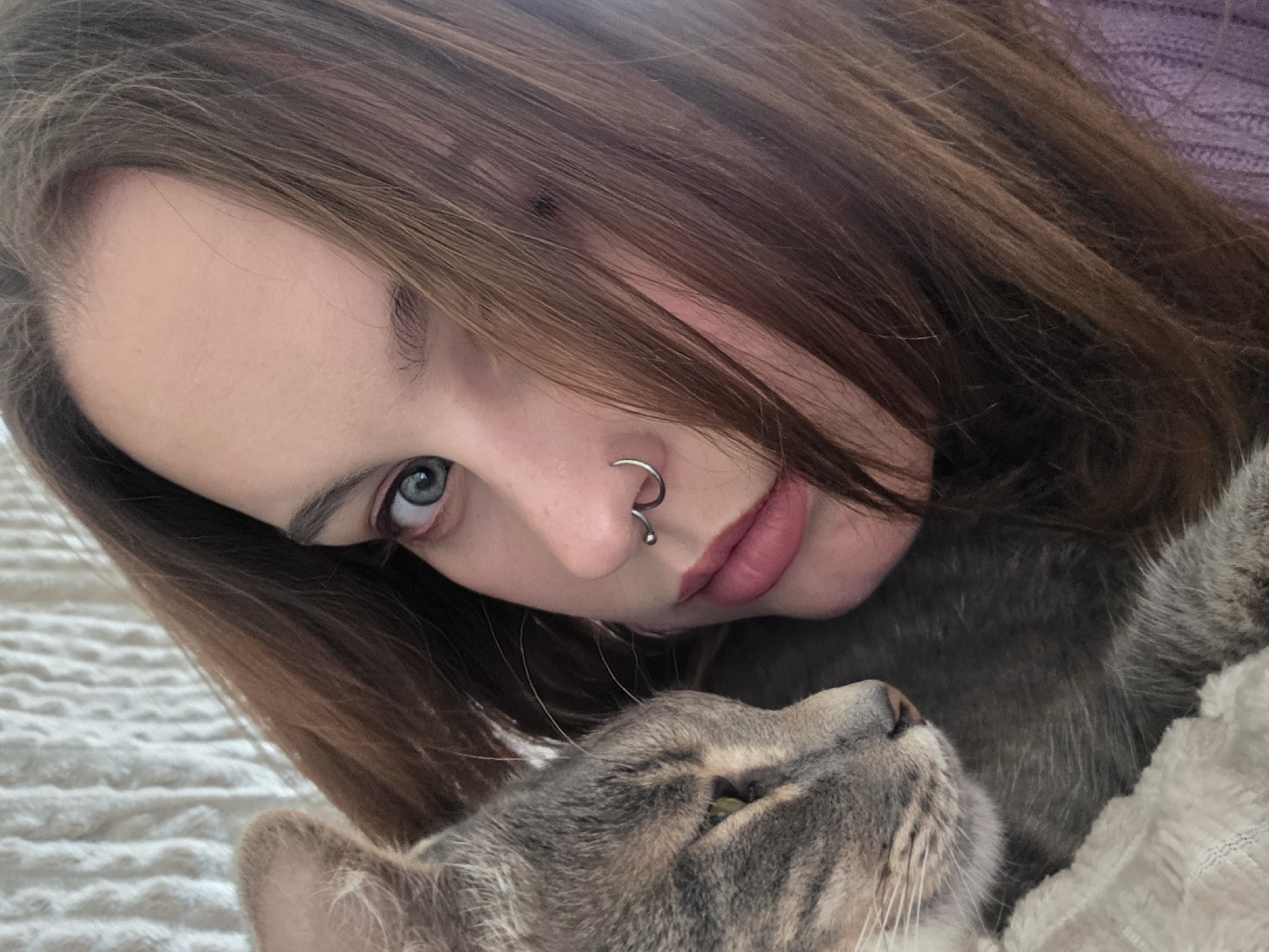A young woman with light skin and brown hair lying on a white textured surface, possibly a bed, is seen in a close-up, angled shot. She is looking toward the camera with her blue eyes. The woman has a silver spiral nose ring on her left nostril. The head of a gray tabby cat is nestled close to the woman's face, with its face turned upwards. The location is unknown.