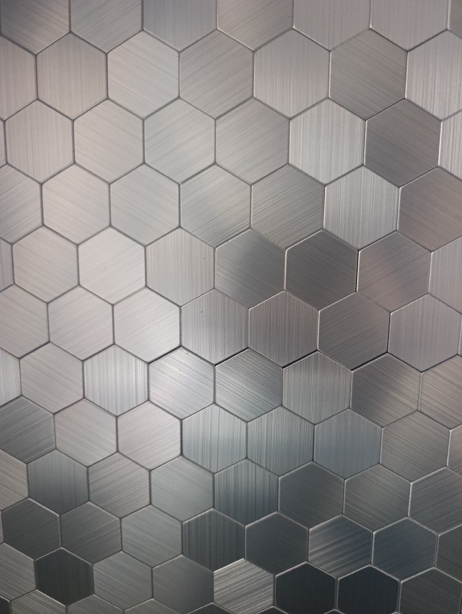 The image shows a close-up, eye-level view of a wall covered in brushed metal hexagonal tiles. The tiles are arranged in a honeycomb pattern, and the brushed texture creates a metallic sheen with visible lines of light reflection. The color palette is predominantly gray and silver, with subtle variations in tone across the tiles due to the lighting and the brushed finish. There are no people or other discernible objects or actions in the image. The background is entirely composed of these tiles. The image does not contain any information that would allow for its location to be identified as Naro-Fominsk, Russia.
