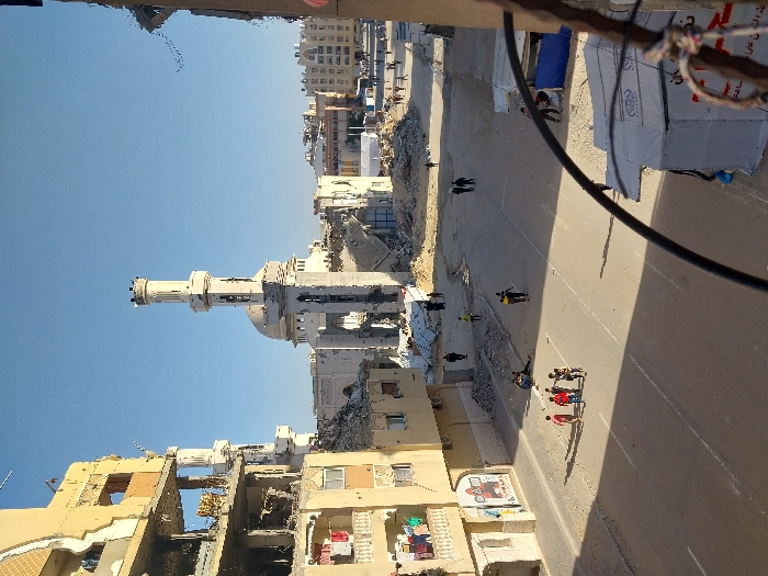 An elevated view captures a severely damaged urban street scene in Khan Yunis, Palestinian Territory, under a clear blue sky. In the center, a large, light-colored mosque features a tall, largely intact minaret; however, its main dome and surrounding structures are significantly destroyed, with large sections collapsed and extensive rubble piled at its base. To the left, a multi-story building exhibits severe structural damage, with outer walls partially missing, exposing interiors and reinforcing bars; laundry hangs on a balcony of this building. Piles of concrete debris, rebar, and general demolition rubble are prominent throughout the scene. An asphalt road traverses the foreground and mid-ground. Approximately five individuals, appearing to be children or young adults, are walking together on the street in the mid-ground, one wearing a red shirt. Additional individuals are sparsely distributed further down the street and near damaged structures in the background. A blue and white makeshift tent or shelter, bearing Arabic text, is visible in the lower right foreground. Strong shadows indicate direct sunlight. A curved black cable or bar partially obstructs the view in the lower right corner.