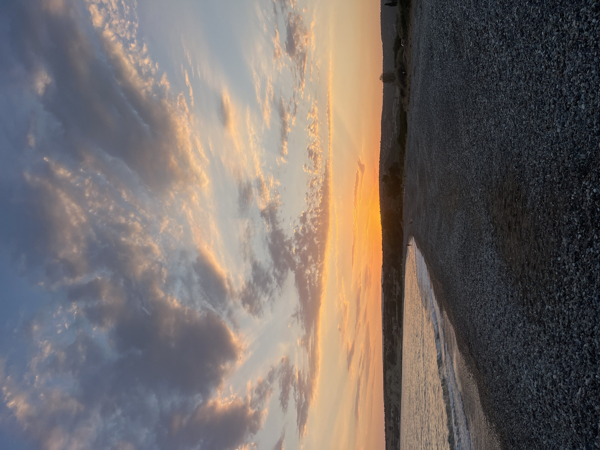 The image depicts a coastal scene at sunset, likely in Avdimou, located within the Sovereign Base Areas of Akrotiri and Dhekelia. The foreground is a dark, pebble-strewn beach with gentle waves lapping at the shore. The sea is a muted silver, reflecting the sky. The middle ground shows a slightly elevated, treed coastline with a few distant figures walking along the beach, their forms silhouetted against the horizon. The sky is a dramatic display of sunset colors, with vibrant oranges and yellows radiating from the setting sun, interspersed with streaks of pink and gray clouds against a soft blue sky. The overall impression is one of tranquility and natural beauty.