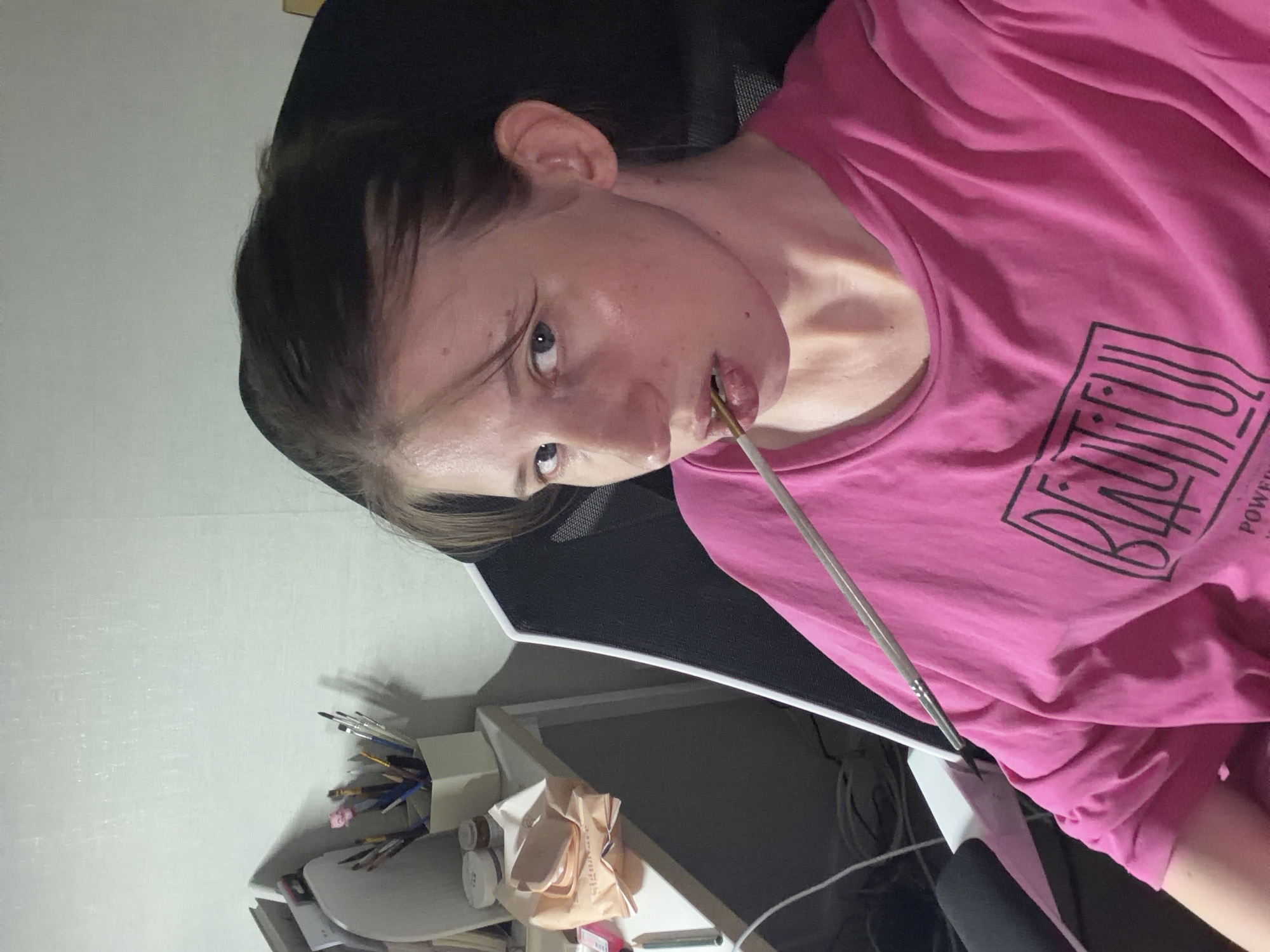 A young woman with light brown hair pulled back is seated in a black gaming chair, wearing a bright pink t-shirt with a graphic print. She is holding a paint brush in her mouth, resting on her lower lip. The woman is looking directly at the camera, her expression neutral. To the left of the chair, a white desk is visible, cluttered with various art supplies, including brushes in a container, jars of paint or other materials, and a package of wet wipes. The background consists of a textured, light-colored wall. The scene appears to be an indoor setting, possibly an artist's studio or a personal workspace, located in Dongducheon-si, South Korea.
