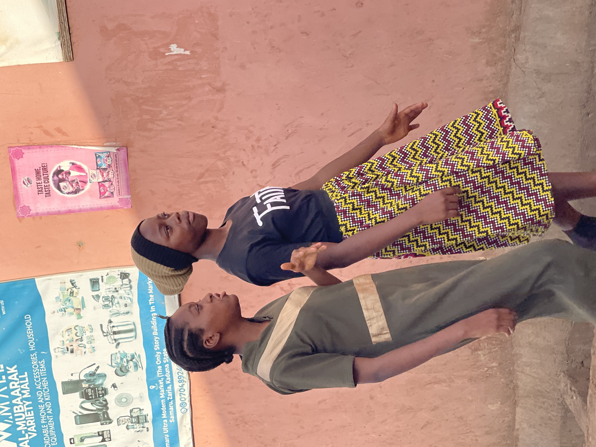 Two young women are standing outdoors in Samaru, Nigeria, captured in what appears to be daytime with natural lighting. They are positioned in front of a textured, reddish-brown wall, possibly the exterior of a building.
The woman on the right wears a black t-shirt with white text (partially visible, possibly "I'M H"), a multi-colored head covering, and a vibrant skirt with a zig-zag pattern in yellow, maroon, black, and white. Her hands are slightly raised, and she looks upwards and to the right. The woman on the left wears a long, olive green dress with light brown or khaki stripes across the chest and shoulders. She has braided hair and also looks upwards and slightly to the right, with a neutral expression.
On the wall behind them are two posters. A pink flyer or poster on the upper left side of the frame features the slogan "TASTE HOME, TASTE CULTURE!" along with images of food or snack products. Below it, a larger blue and white banner for "AL-MUBAARAK VARIETY MALL" is visible. This banner advertises "AFFORDABLE PHONE AND ACCESSORIES, HOUSEHOLD EQUIPMENT AND KITCHEN ITEMS." and provides a location: "Samaru Ultra Modern Market, (The Only Story Building In The Marke Samaru, Zaria, Kaduna State" and a phone number "0007048924". The text "10:09" is also visible, likely part of an item pictured on the banner, possibly a clock or screen. The scene suggests a commercial area, possibly near a market in Samaru.