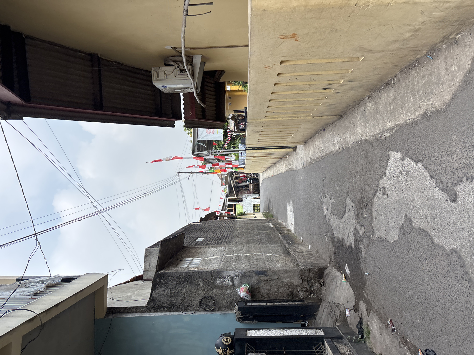 The image captures a narrow, paved alleyway in Bekasi, Indonesia. The alley is lined with buildings on both sides. On the left, a blue-painted building with a white railing on an upper level is visible, adjacent to a rough, grey concrete wall. A black metal gate with a decorative design is also present on the left, with a motorcycle helmet resting on a pillar. The grey wall has a plastic bag filled with items hanging from it.
On the right side of the alley, a light yellow building with a corrugated metal roof overhang dominates the view. An outdoor air conditioning unit is mounted on the wall of this building. A low, yellow-painted concrete wall with vertical rectangular openings runs along the edge of the alley.
The alley itself appears weathered, with patches of water and debris on the asphalt. Further down the alley, Indonesian flags, red and white, are strung across. Several stalls or small shops are visible in the distance, with some laundry hanging out to dry. The sky is overcast with scattered clouds. The scene suggests a residential or mixed-use area within the city.