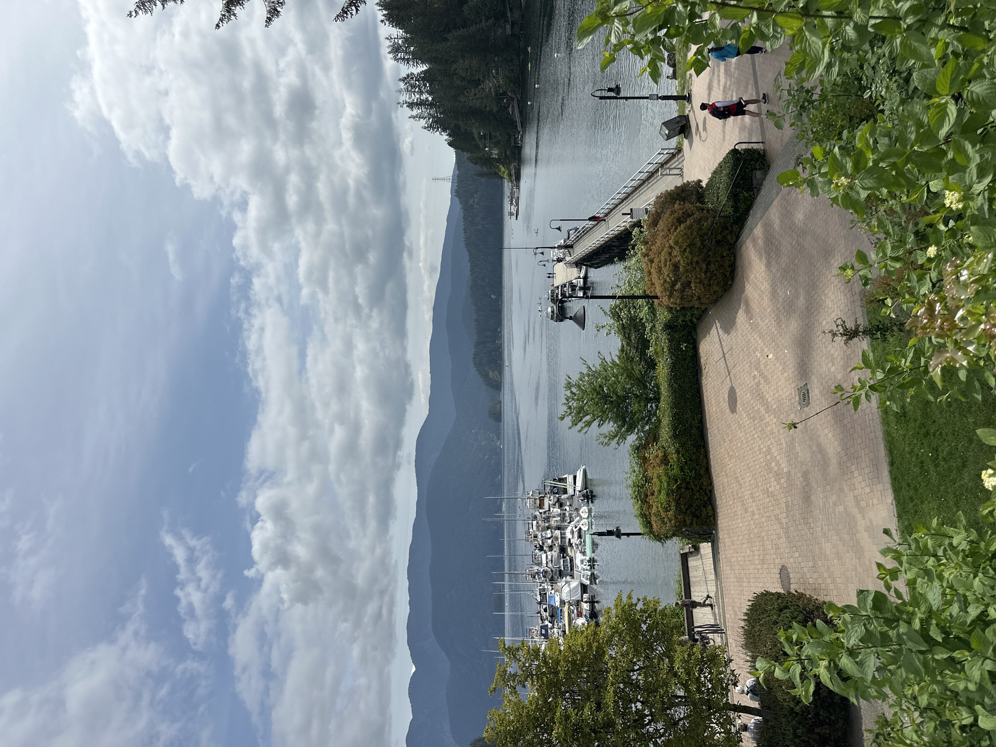 This image captures a picturesque scene in the District of North Vancouver, Canada, showing a harbor filled with boats and yachts. A paved walkway lined with bushes and trees runs along the water's edge, where people can be seen strolling. The backdrop features a range of mountains under a sky filled with clouds, adding depth to the scenery. A pier extends into the water, further enhancing the harbor's charm.