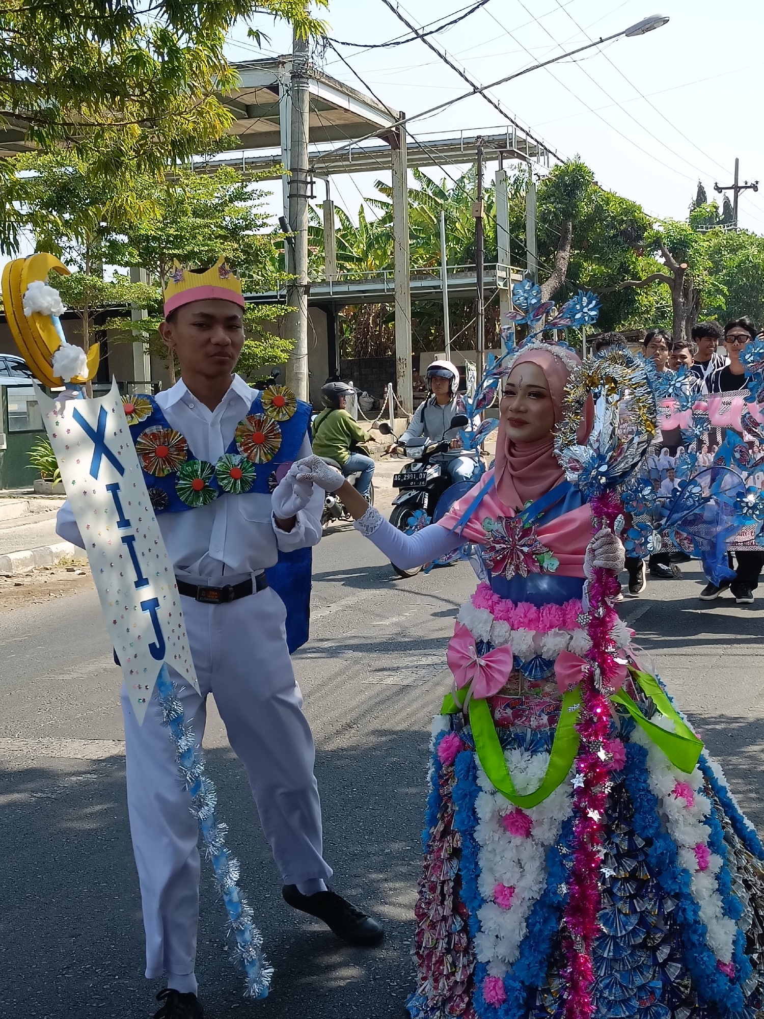 Two individuals are participating in a parade on a street in Kediri City, Indonesia. The person on the left, a male, wears a white long-sleeved shirt, white pants, and a pink and yellow paper crown. He holds a long, white banner adorned with colorful confetti and the letters "XII IJ" in blue. He also holds a yellow crescent moon prop with white cotton clouds attached. Around his neck, he wears a blue sash decorated with circular, colorful ornaments. The person on the right, a female, is dressed in an elaborate costume. She wears a pink hijab and a pink and blue dress that is very full and adorned with many colorful embellishments, including bows, tissue paper flowers, and what appear to be candy wrappers. She holds a tall, decorative staff. Both individuals are wearing white gloves. In the background, there are trees, a street lamp, power lines, construction scaffolding, and other people, some of whom are on motorcycles. The overall scene suggests a festive cultural event.