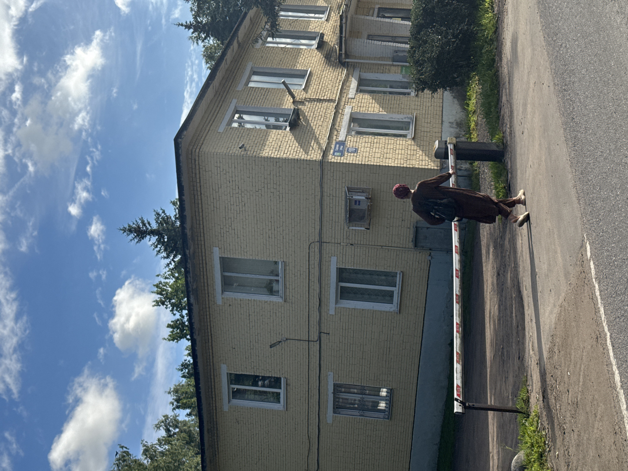In Annino, Russia, a person wearing a brown robe and head covering walks past a yellow brick apartment building. The building has several windows with white frames, and a street sign reading "Sadovaya ulitsa 10" is visible on the exterior wall. A white and red striped barrier extends across the foreground. The sky is blue with scattered white clouds, and trees are visible behind the building.