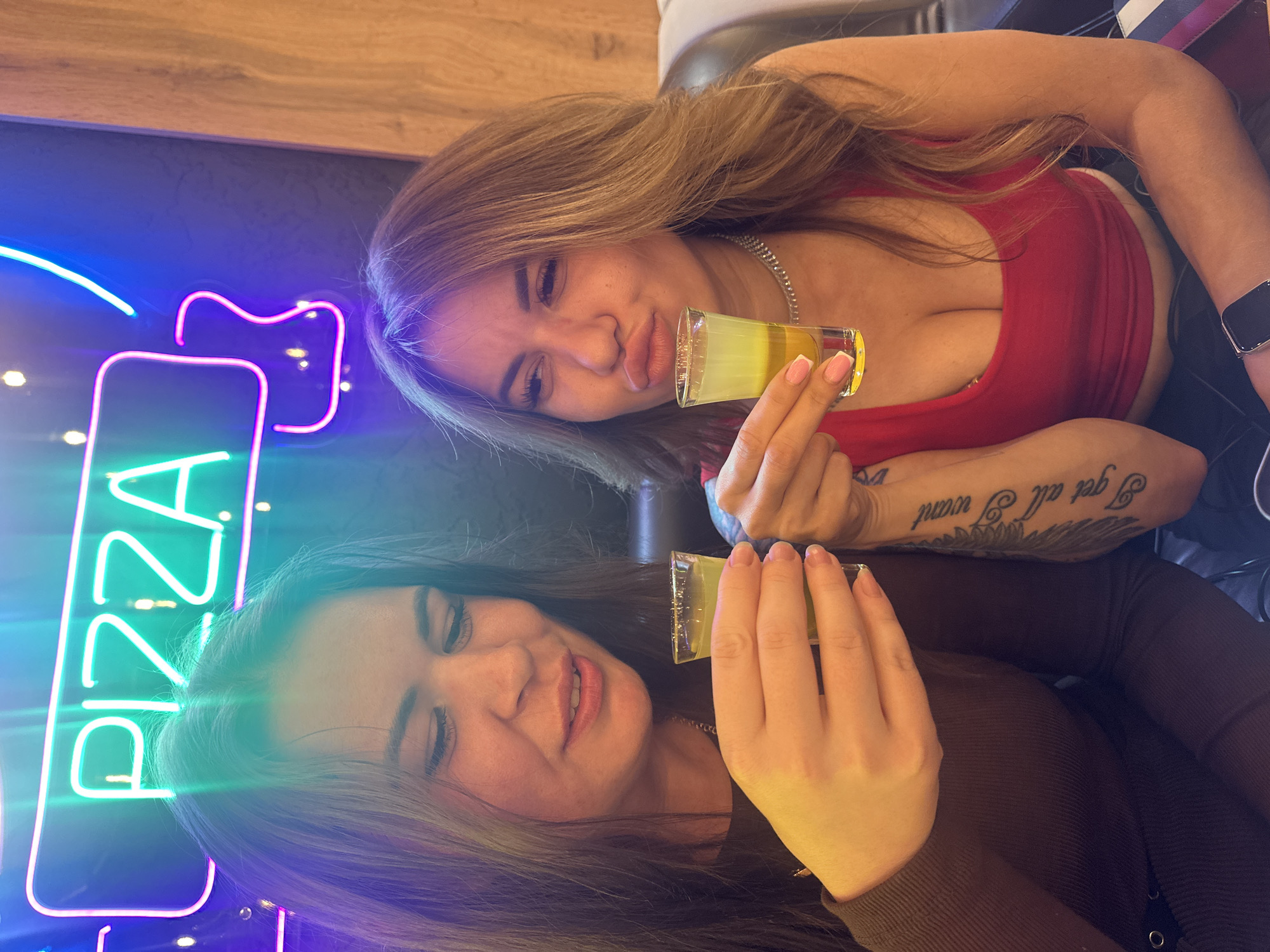 Two women are posing indoors with small glasses of liquid, possibly alcoholic beverages. A neon sign with the word "PIZZA" is visible on the left. The woman at the top has light brown hair and is wearing a red top. The woman below has dark hair and is wearing a brown top. The exact location and its relation to Perm, Russia, are not discernible from the image alone.
