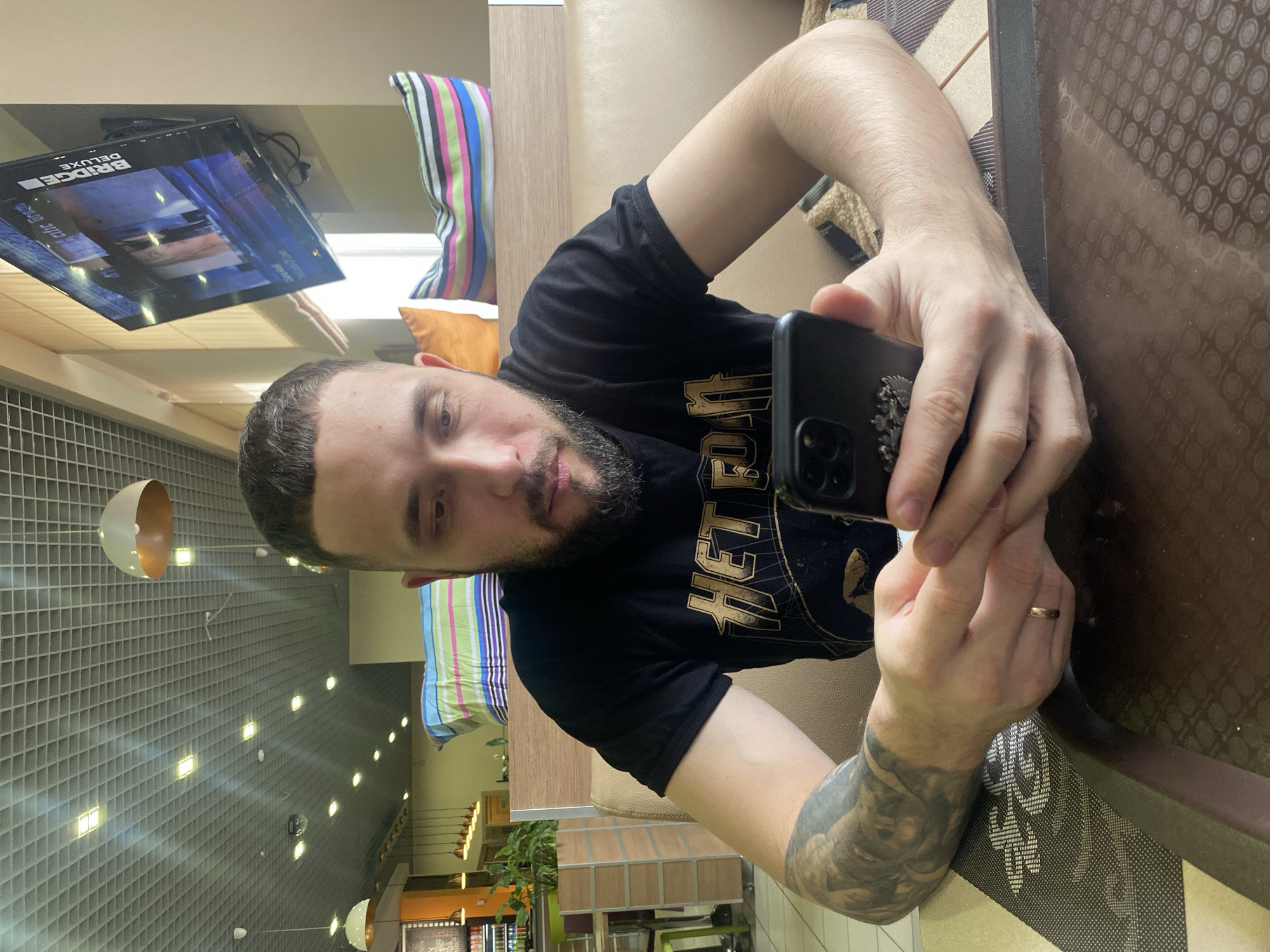 A medium shot depicts a man sitting at a table and holding a smartphone. He is wearing a black t-shirt with gold lettering. His left arm, visible to the viewer, has a detailed tattoo. He has dark hair and a beard. He appears to be taking a photo or video with his phone. The table has a brown, patterned surface. In the background, there's seating with colorful pillows. A television screen showing "BRIDGE DELUXE" is mounted on the ceiling. The location is in the city of Chaikovsky, Russia, as suggested by the prompt.