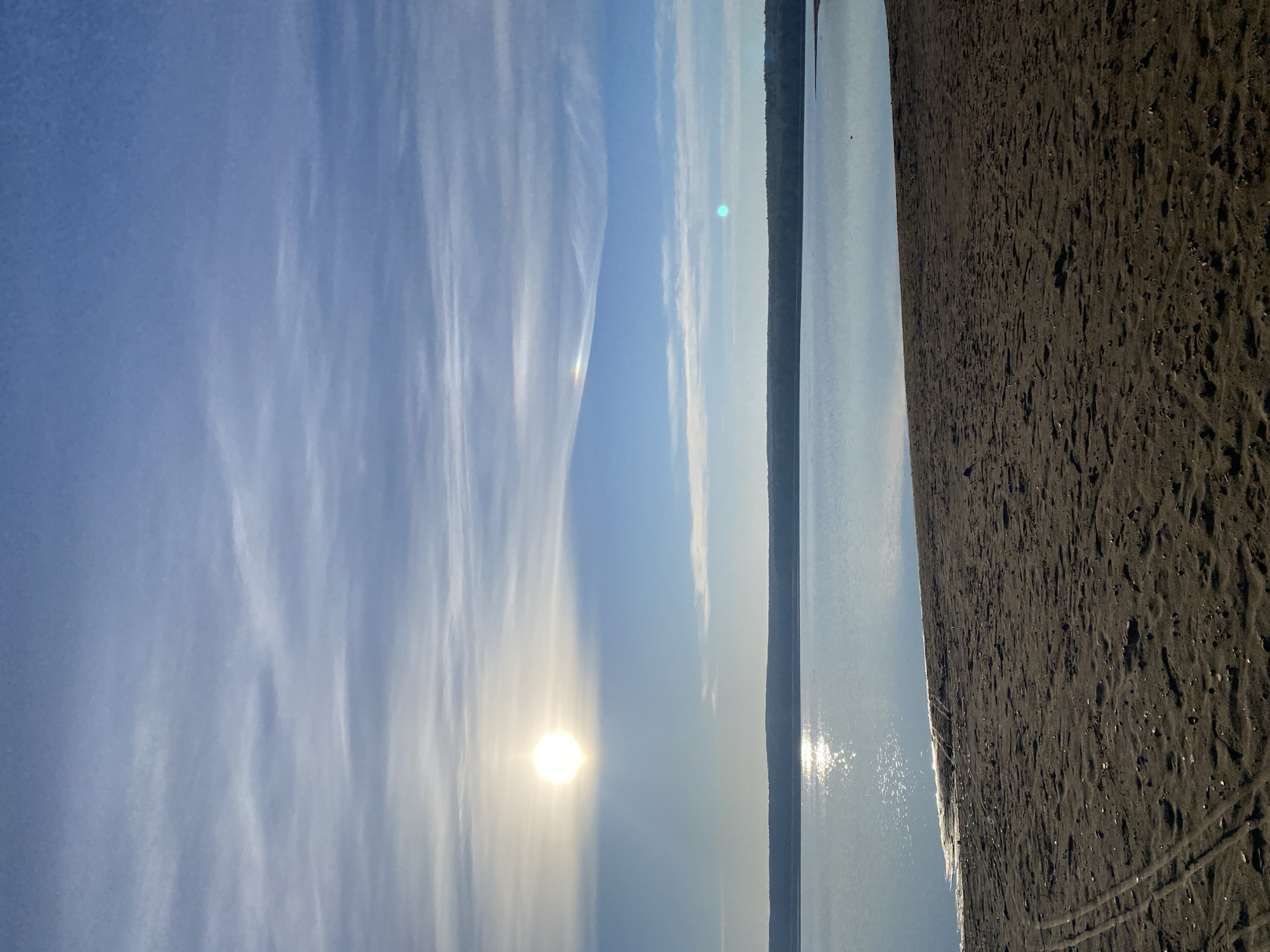 The image depicts a landscape at the shore, likely in Chaikovsky, Russia. The scene includes a body of water, possibly the Kama River, with the sun reflecting off its surface. The sky is blue with cirrus clouds, and the sun is visible. To the right is a sandy beach with traces of tracks on the sand. Vegetation or land is visible in the distance across the water. There is no indication of the presence of people.