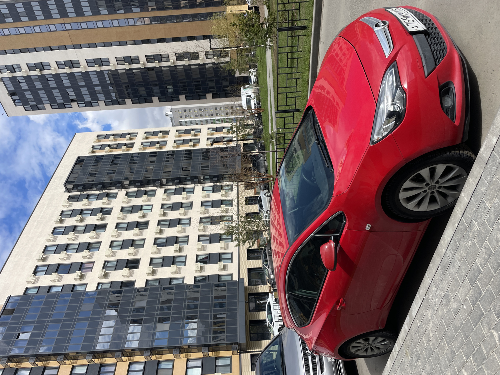 The image shows a red Opel Astra parked on a paved street in Kazan, Russia. The license plate is A 759MK 116. In the background are several multi-story residential buildings with a mix of light-colored facades, dark glass paneling, and numerous air conditioning units. The sky is partially cloudy. A metal fence runs along the street near the sidewalk. There are other parked cars behind the red Opel.