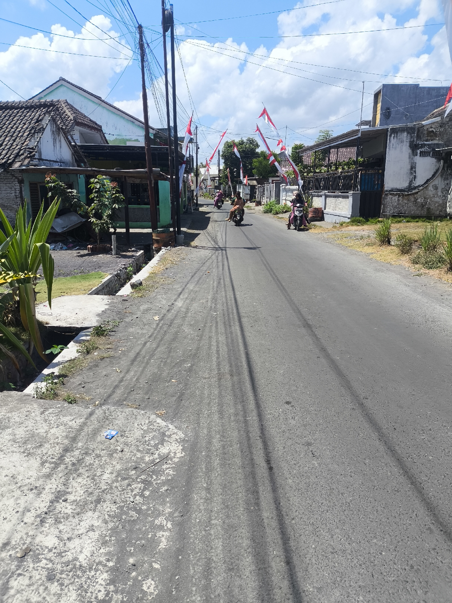 This is a street scene in Kebonsari, Indonesia, on a sunny day with scattered clouds. The street is paved with asphalt and appears to be a residential area, with houses on either side. Red and white Indonesian flags are strung across the street, suggesting a national celebration. Several people are riding motorcycles on the street. On the left side of the street, there is a building with a tiled roof and a green facade. There is also a large plant with long green leaves in the foreground. On the right side of the street, there are more houses and dry grass. There are several utility poles with numerous electrical wires crisscrossing the sky. A small, blue, crumpled piece of plastic is visible on a concrete surface in the lower left quadrant of the image.