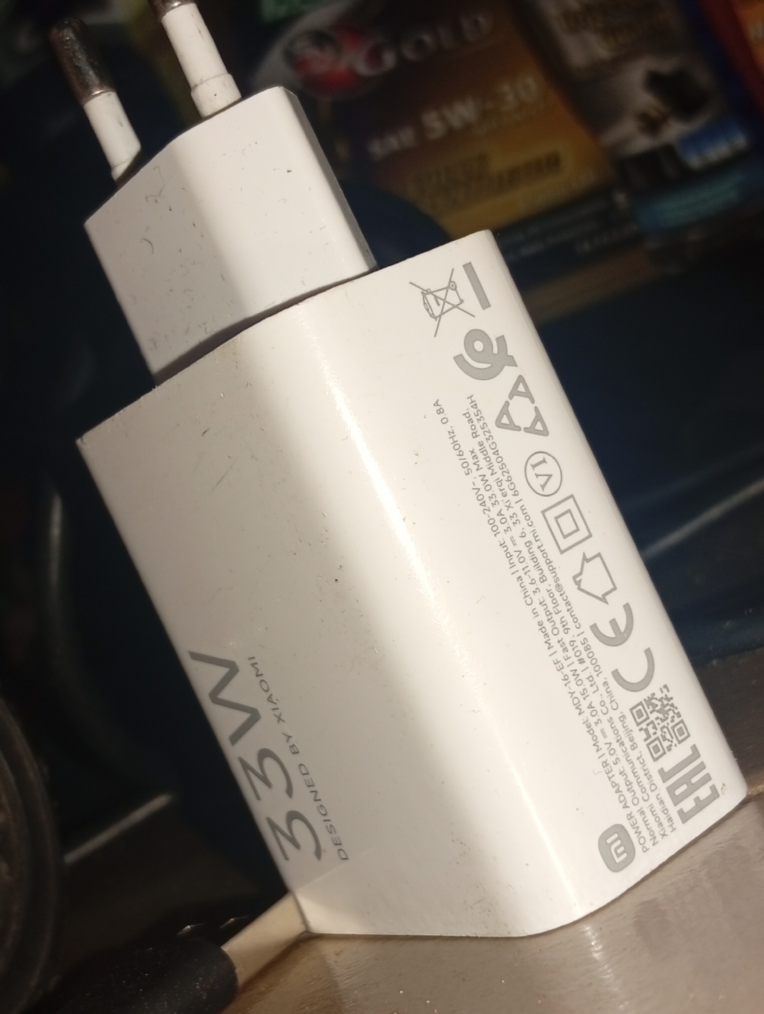 A white Xiaomi 33W fast charger is shown in the image. The charger has visible dust and smudges on its surface. The text "33W DESIGNED BY XIAOMI" is printed on the charger in a gray font. Other markings on the charger include various logos and technical specifications such as "Input: 100-240V~ 50/60HZ 0.8A" and "Output: 5V 3.0A 15.0W; 9V 2.23A 20.07W; 12V 1.67A 20.04W". The charger is positioned at an angle, with its European-style prongs pointing upwards. The background is dark and out of focus, showing some blurry items that appear to be other electronic devices or packaging. There is no indication of the location being Ukukwa, Nigeria.