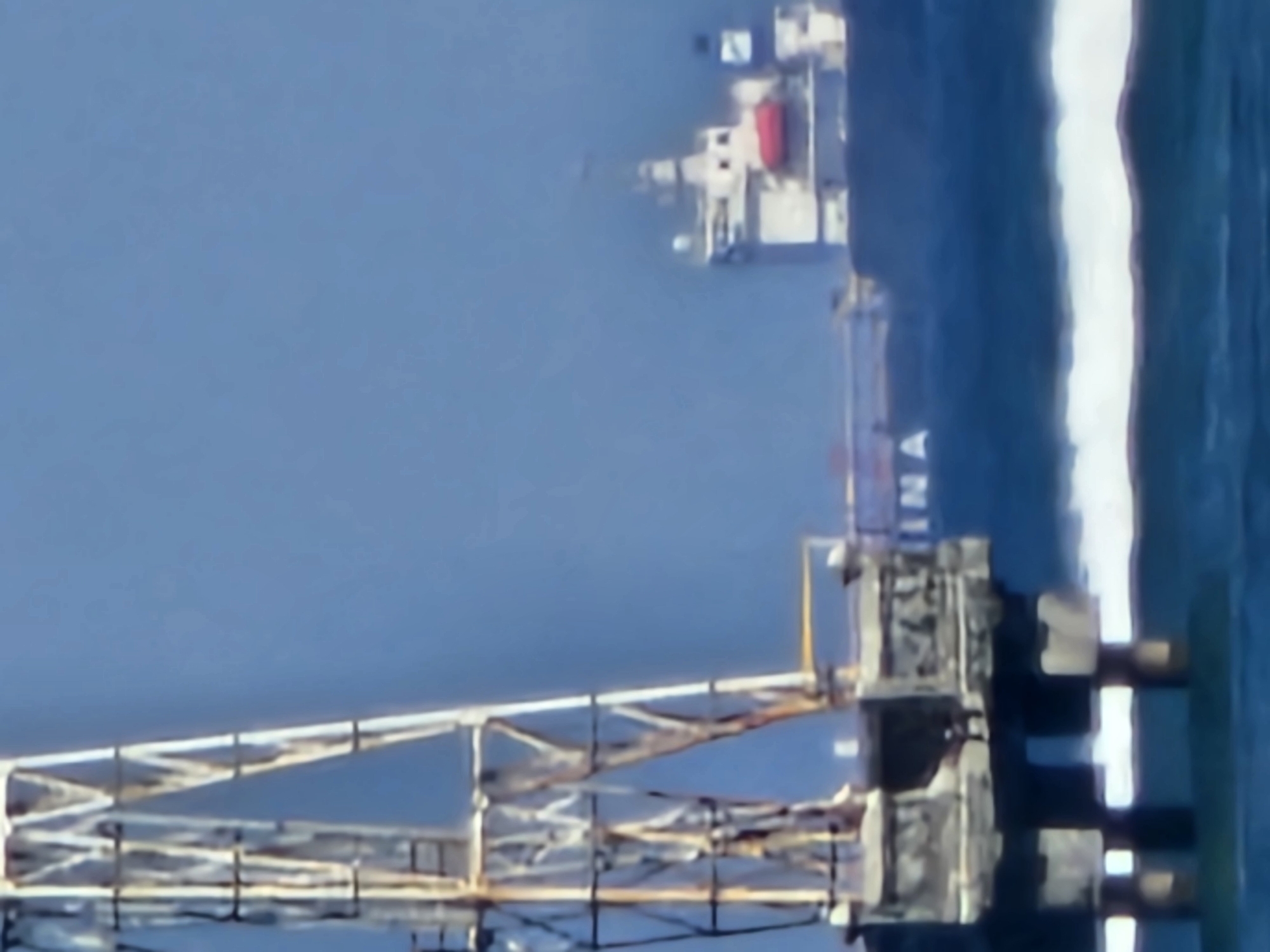 An offshore platform with a metal structure is visible in the foreground on the left. In the background, a large ship with a white superstructure and a red life raft is sailing on the water. The sky is a clear blue. The scene is likely a view from a coastal area, possibly Denpasar, Indonesia, known for its beaches and maritime activities.
