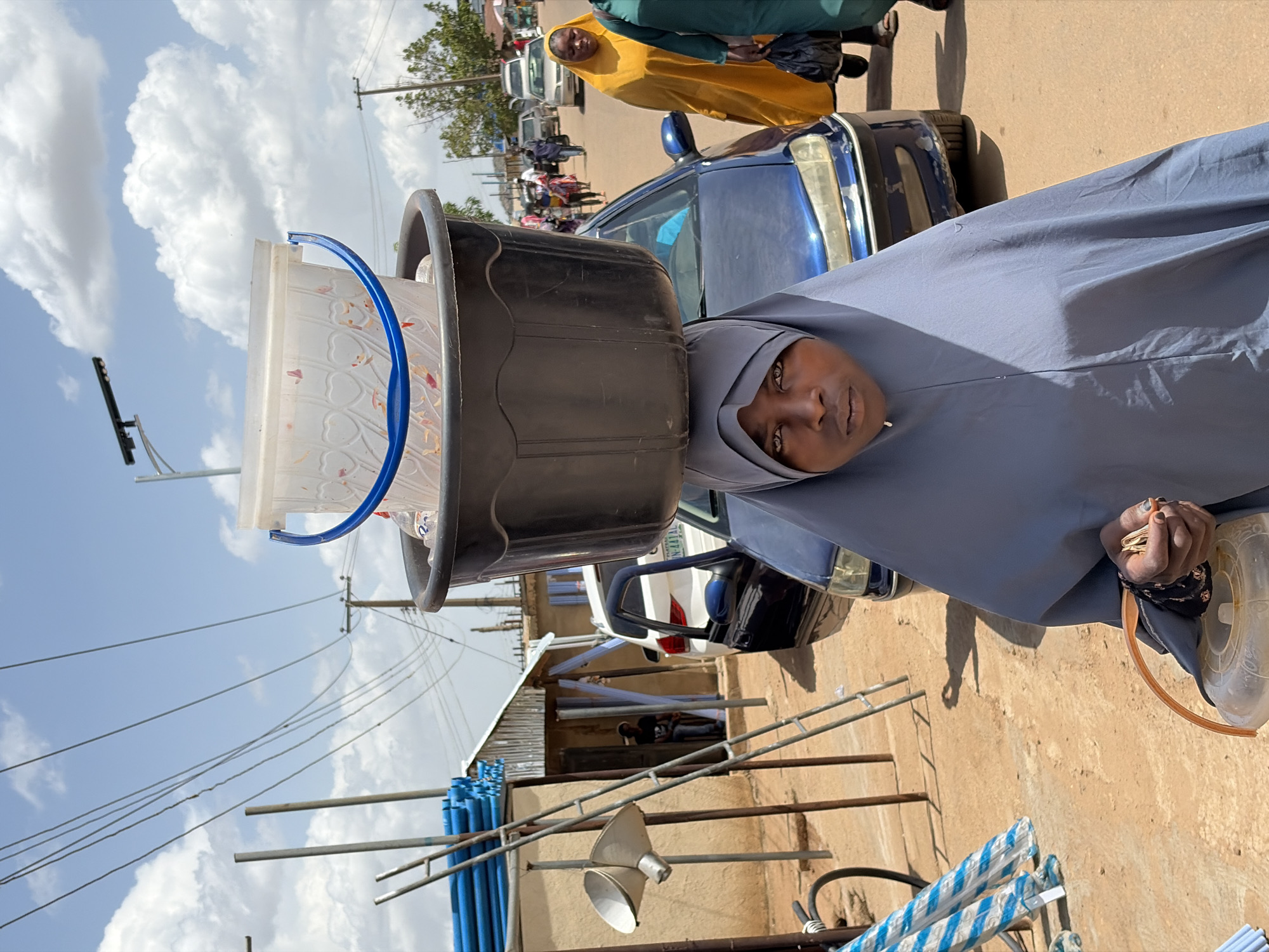 This vertical-format image captures a street scene in Gura Top, Nigeria, featuring a woman balancing containers on her head. The main subject, positioned on the right side of the frame, is a dark-skinned woman wearing a grey hijab that covers her head and shoulders, along with a long, flowing grey garment. She looks directly at the camera with a neutral expression. On her head, she skillfully balances a large, black, possibly plastic, bucket, on top of which sits a clear plastic container with a distinctive blue handle. Inside the clear container, small, colorful items are visible, possibly confectionery or snacks for sale. In her right hand, she holds a handful of small, light-colored objects, resembling nuts or seeds, and a flat, brown, disc-shaped item. Her fingernails are painted dark.
In the mid-ground, behind the woman, a dark blue sedan is parked or moving, partially visible. Further back and to the left, another person in a bright yellow head covering and garment is visible, also looking towards the camera. The background reveals a bustling street environment with other vehicles and figures. The ground is unpaved, appearing dusty and brown.
Above, power lines stretch across a clear blue sky dotted with scattered white clouds. Utility poles are visible in the distance. To the left, structures with corrugated metal roofs and stacks of blue pipes or rods suggest ongoing construction or a commercial area. A silver, funnel-shaped object rests near the bottom left. The bright lighting indicates it is daytime, likely mid-day, with strong shadows present. A blurry license plate on a white vehicle in the background contains text that is unreadable in the main image but hinted to be "NIGERIA" in cropped views.