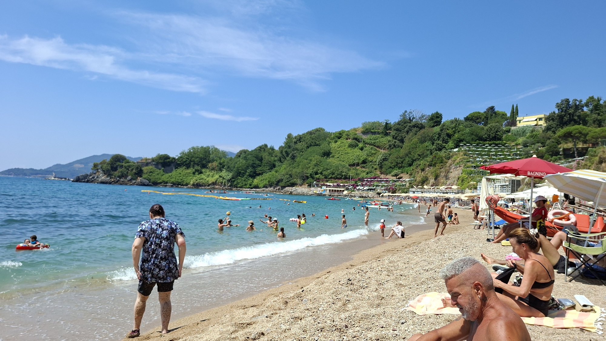 This is a daytime, eye-level, wide shot of a crowded beach scene in Lerici, Italy. The beach is lined with sunbathers and swimmers enjoying the clear blue water and sunny sky. In the foreground, a man with grey hair is sitting on a towel, looking to the left. To his right, a woman is also sitting on a towel, wearing a black bikini and looking down. Behind them, several beach umbrellas and chairs are set up, with a red umbrella prominently displayed with the word "BAGNINO" on it, indicating a lifeguard station.
The midground shows a lively beach with many people. Some are swimming in the shallow water, while others are playing with a volleyball. Yellow buoys mark off a swimming area. Further out in the water, several small boats and inflatable toys are visible. The background features a steep, green, tree-covered hillside with buildings and more beach umbrellas clustered at its base. The coastline extends to the left, with a rocky outcrop and hills in the distance. The overall impression is of a vibrant and popular summer beach destination.
